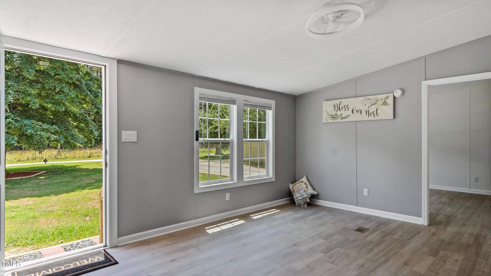 A bright room with light wood flooring, gray walls, two large windows, an open door showing green grass and trees outside, and a decorative sign on the wall reading "bless our nest.
