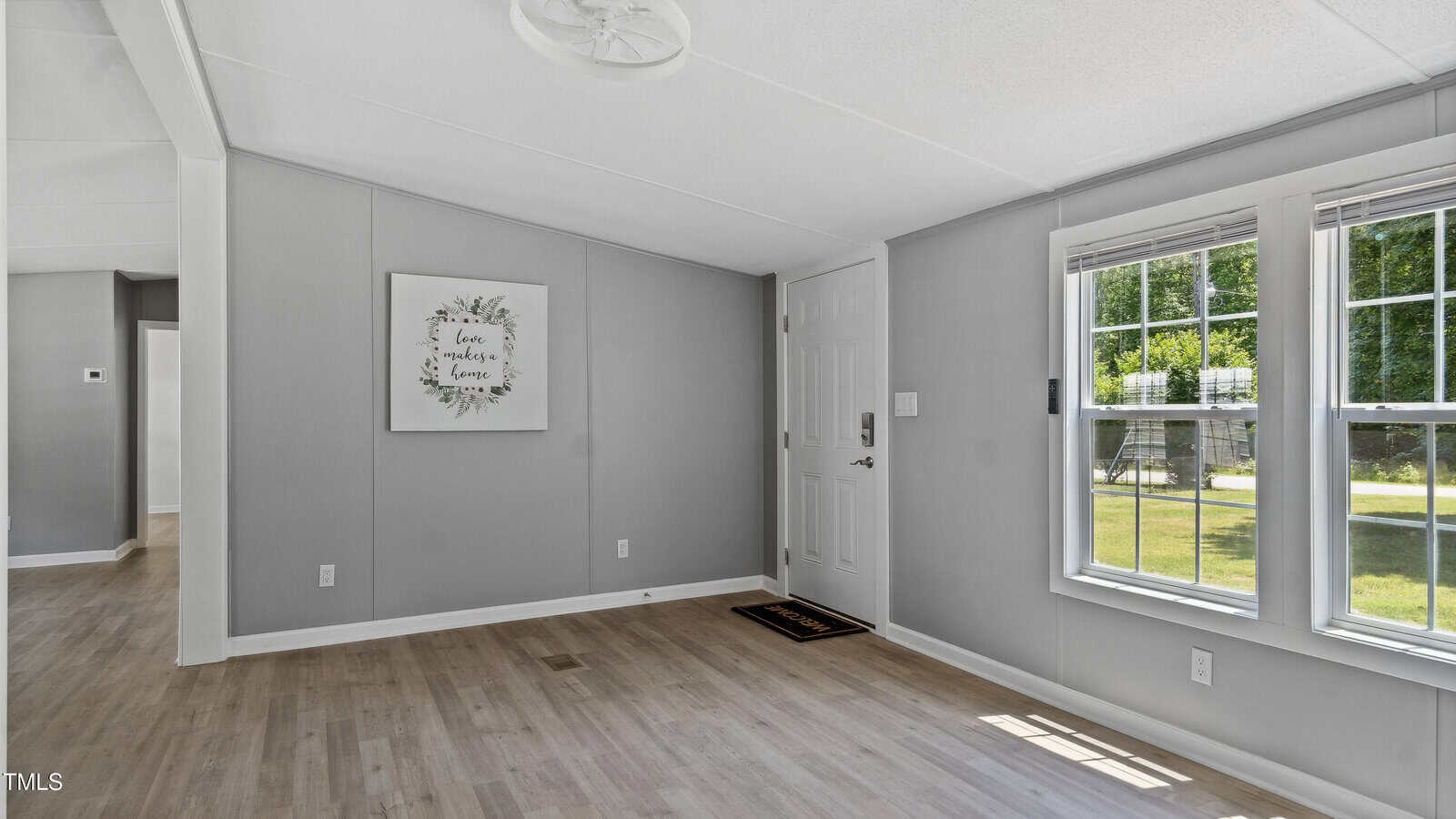 A bright, unfurnished room with light wood floors, gray walls, a white entry door, large windows letting in sunlight, and a framed sign on the wall that reads "Love makes a house a home.