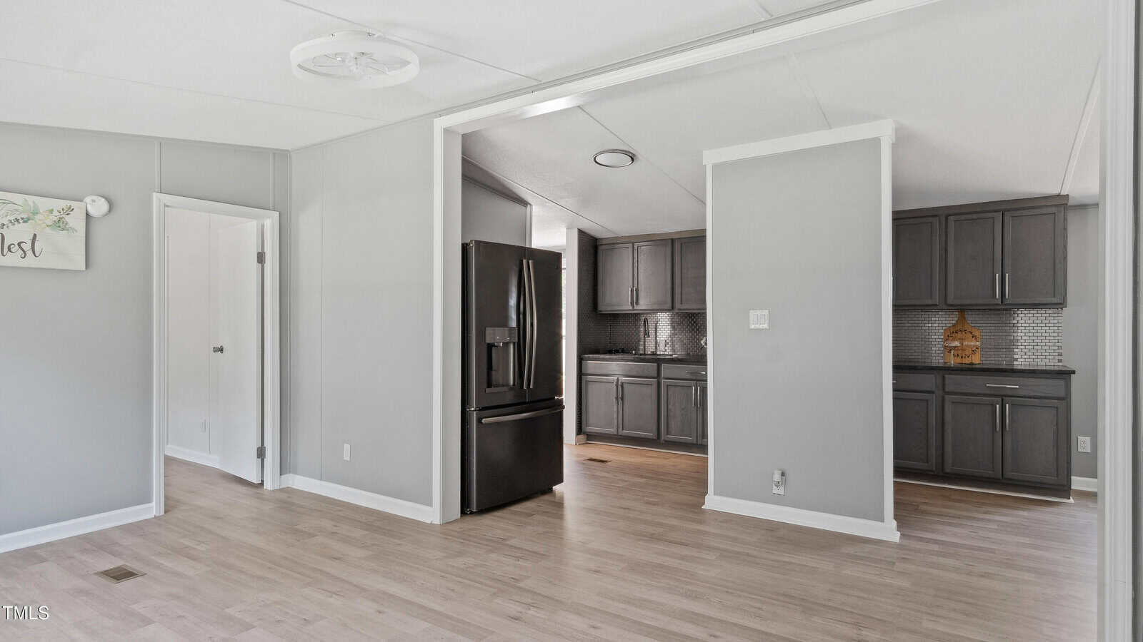 A modern kitchen with dark cabinets, a black refrigerator, light wood floors, and light gray walls. A doorway leads to another room, and there is a decorative sign on one wall.