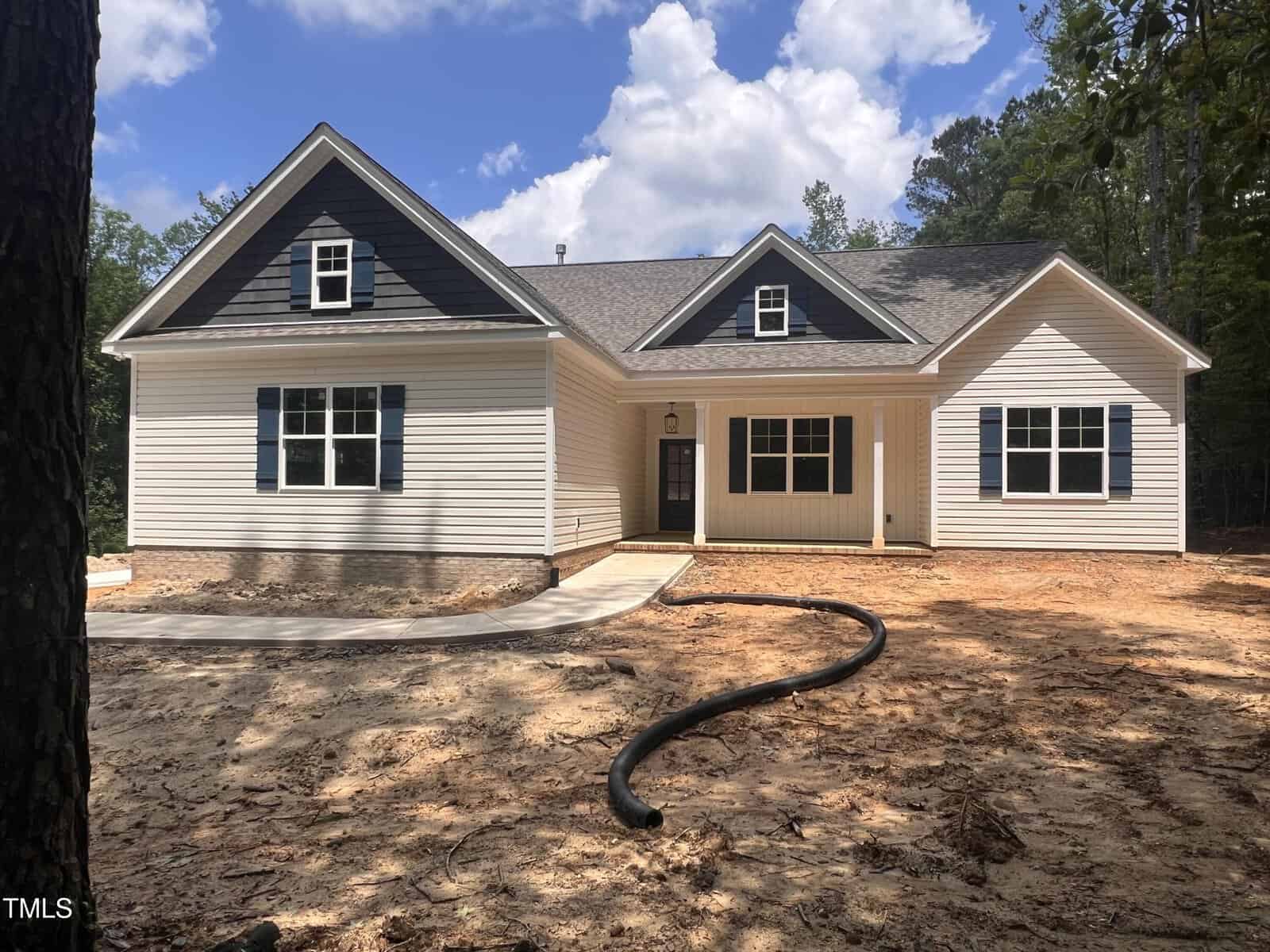 Single-story house with cream-colored siding and dark shutters, a front porch with columns, and a gable roof, set on a dirt lot with surrounding trees and a partly cloudy sky.