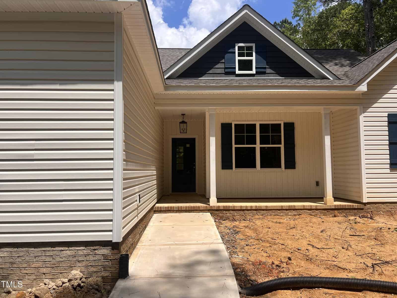 A newly built house with light-colored siding, dark accent shingles on the upper front, a covered entryway, white trim, and a concrete walkway leading to the front door. The yard is unfinished, with exposed dirt.