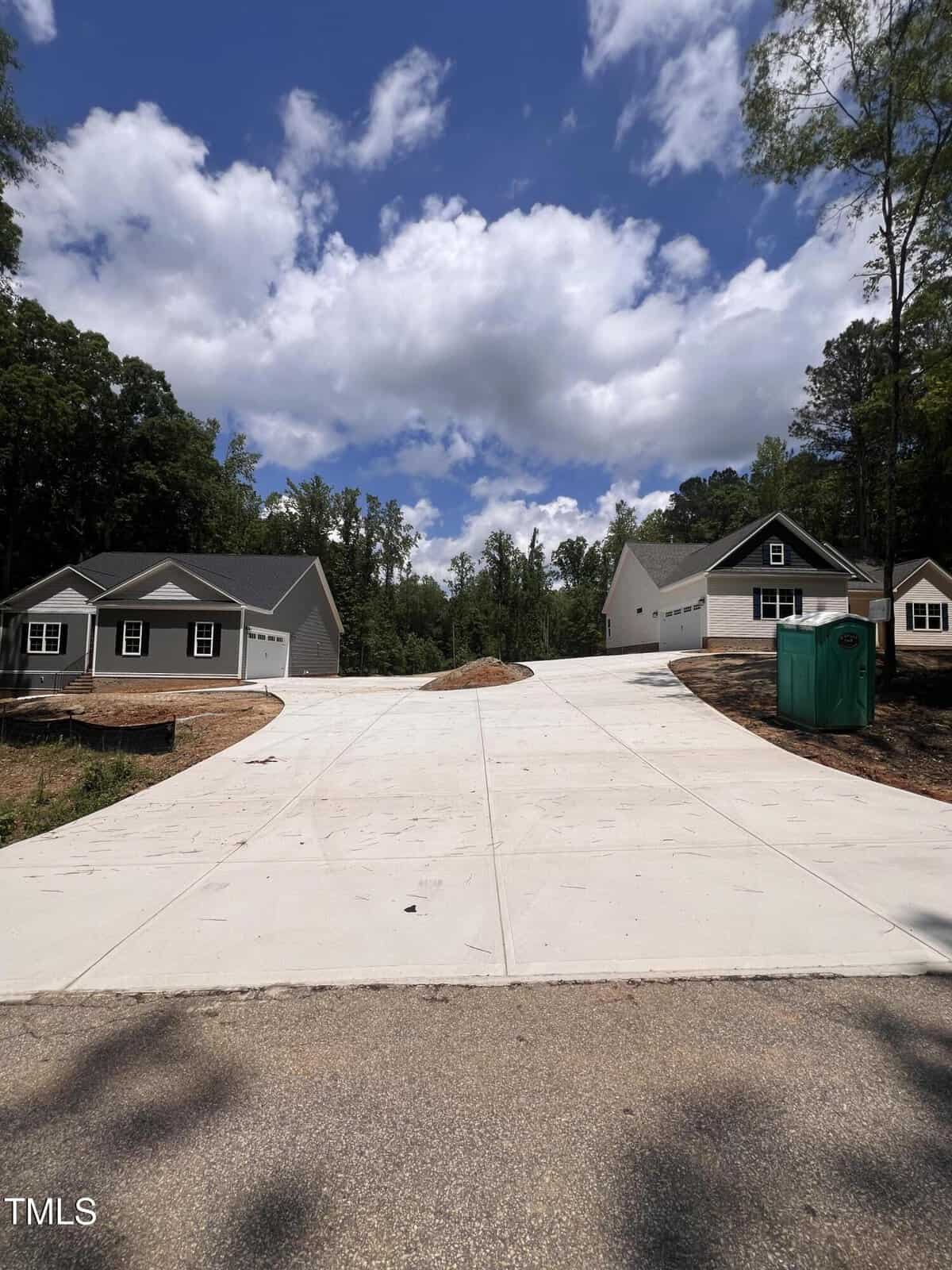 A wide concrete driveway splits into two, leading to two single-story houses with garages on either side. Trees and a green portable toilet are visible. The sky is partly cloudy.