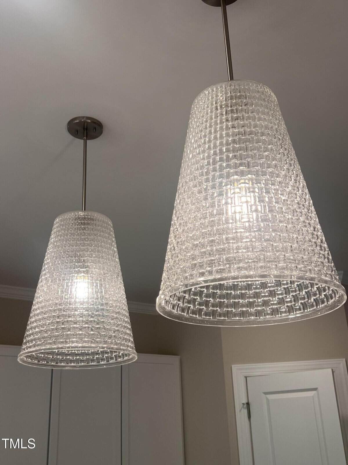 Two pendant lights with textured, translucent glass shades hang from the ceiling in a modern, neutral-toned room. A white door and part of a cabinet are visible in the background.