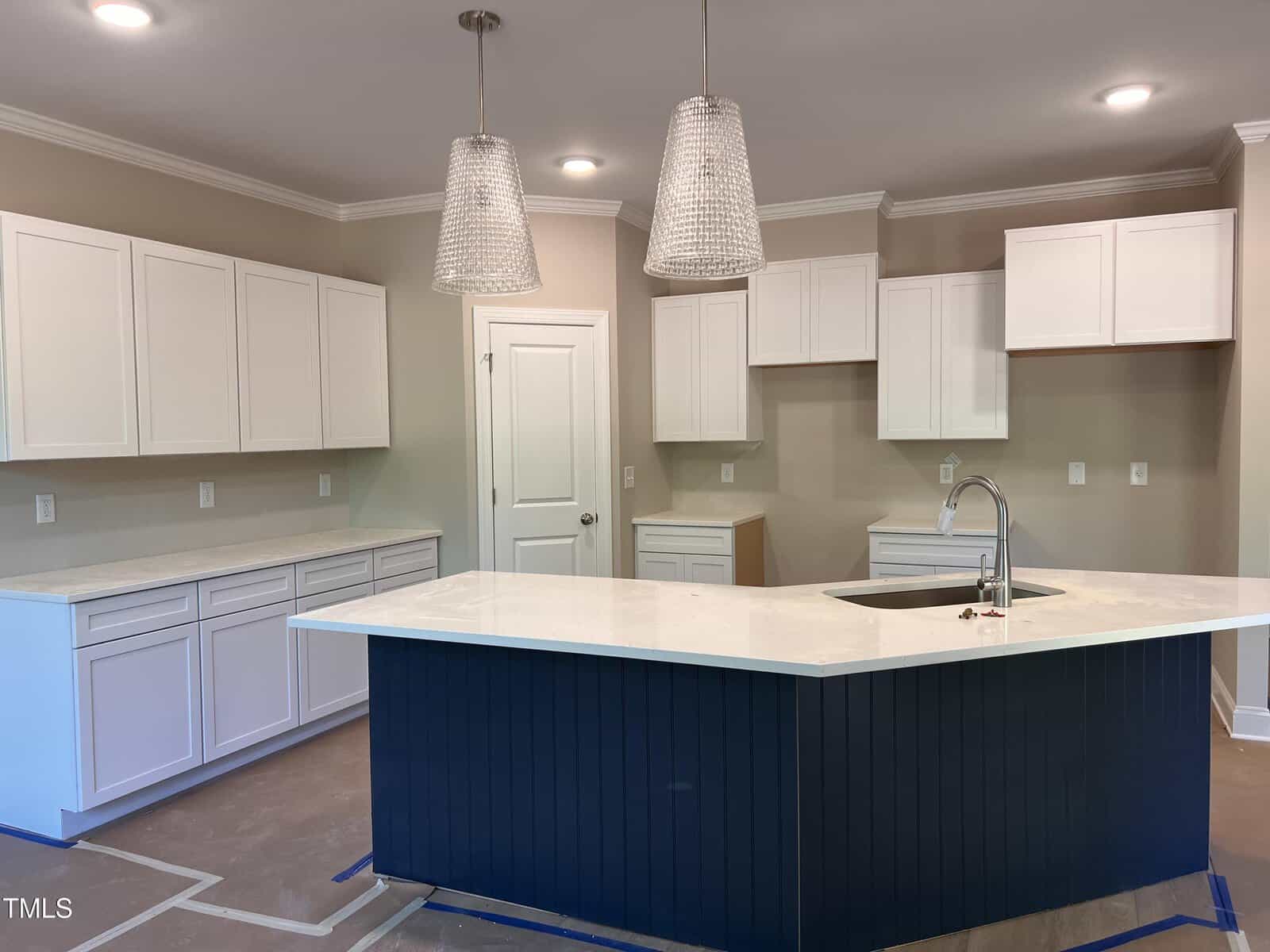 Modern kitchen with white upper and lower cabinets, beige walls, a large dark blue island with a sink, two pendant lights above, and recessed lighting in the ceiling. Countertops are white; floor appears unfinished.