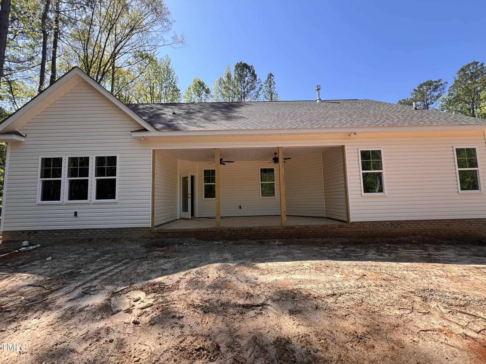 A single-story house with light-colored siding, a large covered back patio supported by wooden posts, ceiling fans, multiple windows, and a dirt backyard, surrounded by tall trees under a clear blue sky.