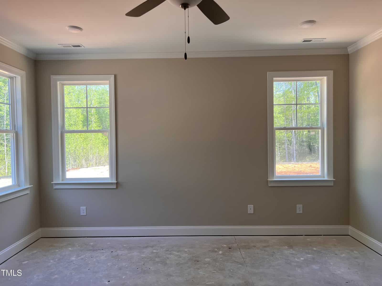 Empty room with light beige walls, two windows on each side wall, and one window on the back wall. Ceiling fan centered above, bare floor with no carpeting or furniture. Natural light comes through the windows.