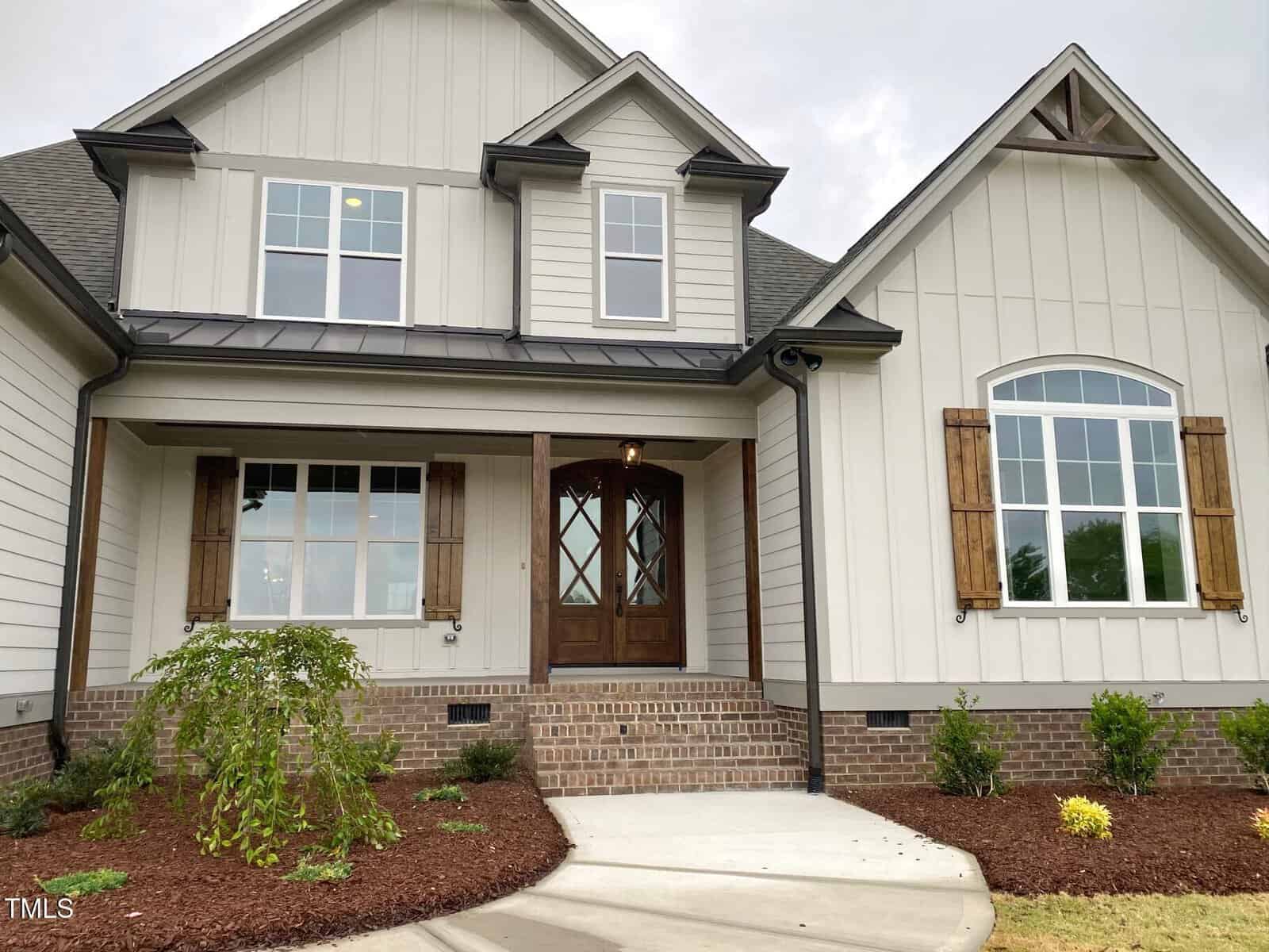 A modern house with light-colored vertical siding, brick foundation, wooden double front doors, and windows accented with brown shutters. There is a small landscaped area and a concrete walkway leading to the entrance.
