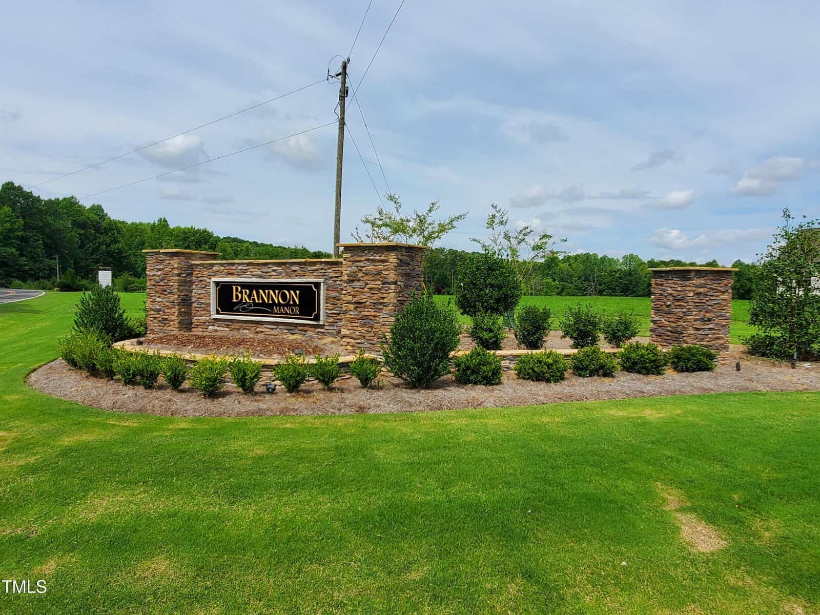 A stone entrance sign reads “Brannon Manor,” surrounded by small bushes and mulch, set on a well-maintained grassy lawn with trees and a road in the background. A utility pole stands behind the sign.