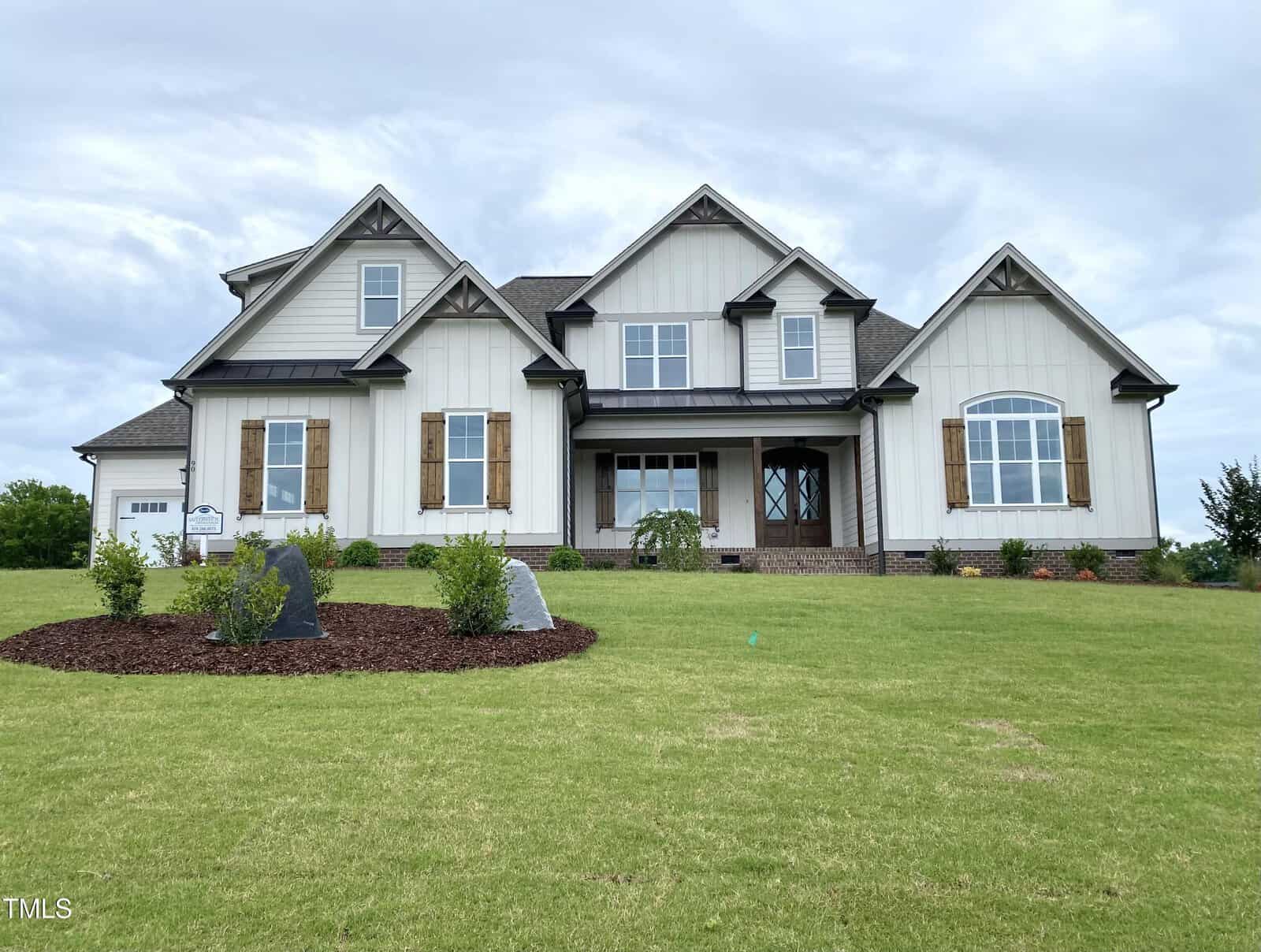 Two-story house with white siding, dark roof, and wooden shutters located at 90 Water Willow Lane in Zebulon. The house features multiple gables and large windows. There is a green lawn with landscaping and shrubs in front of the house under a cloudy sky.