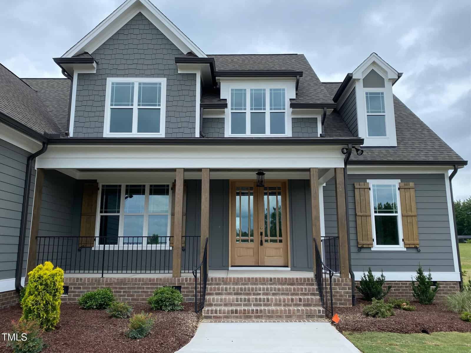 A two-story house with gray siding, white trim, wood accents, a covered front porch with brick steps, double front doors, and several windows. There is simple landscaping with shrubs and a small tree.