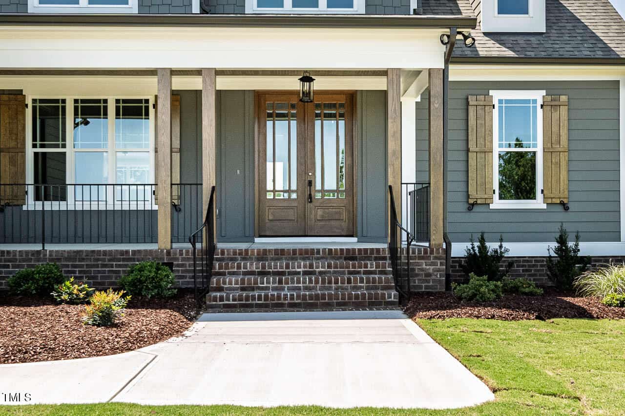 A front porch with brick steps leads to double wooden doors with glass panels. The house exterior is gray with white trim and features a window with wooden shutters and landscaped plants near the entrance.