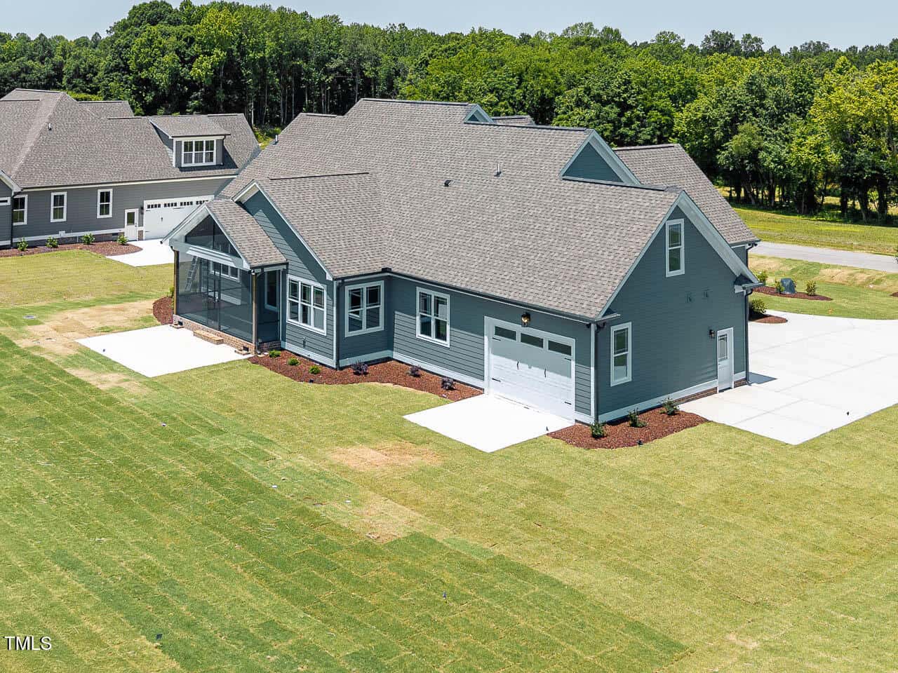 A large, modern blue-gray house with a steep roof, attached garage, screened-in porch, and neatly manicured lawn, situated in a spacious suburban neighborhood with green trees in the background.