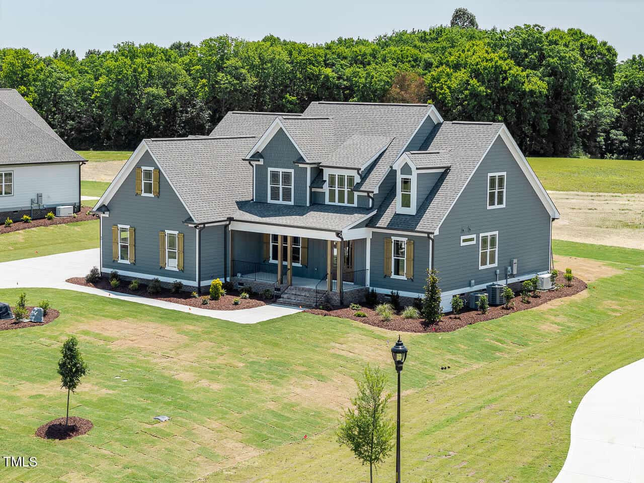 A large, two-story house with gray siding, white trim, and a covered front porch sits on a spacious lot with a lawn, young trees, and a curved driveway. Trees line the background behind the property.