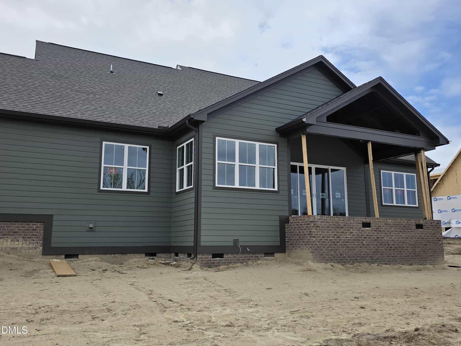 A newly constructed house with green horizontal siding, multiple white-framed windows, a gabled roof, and a covered back porch with brick columns. The yard is bare dirt with no landscaping.