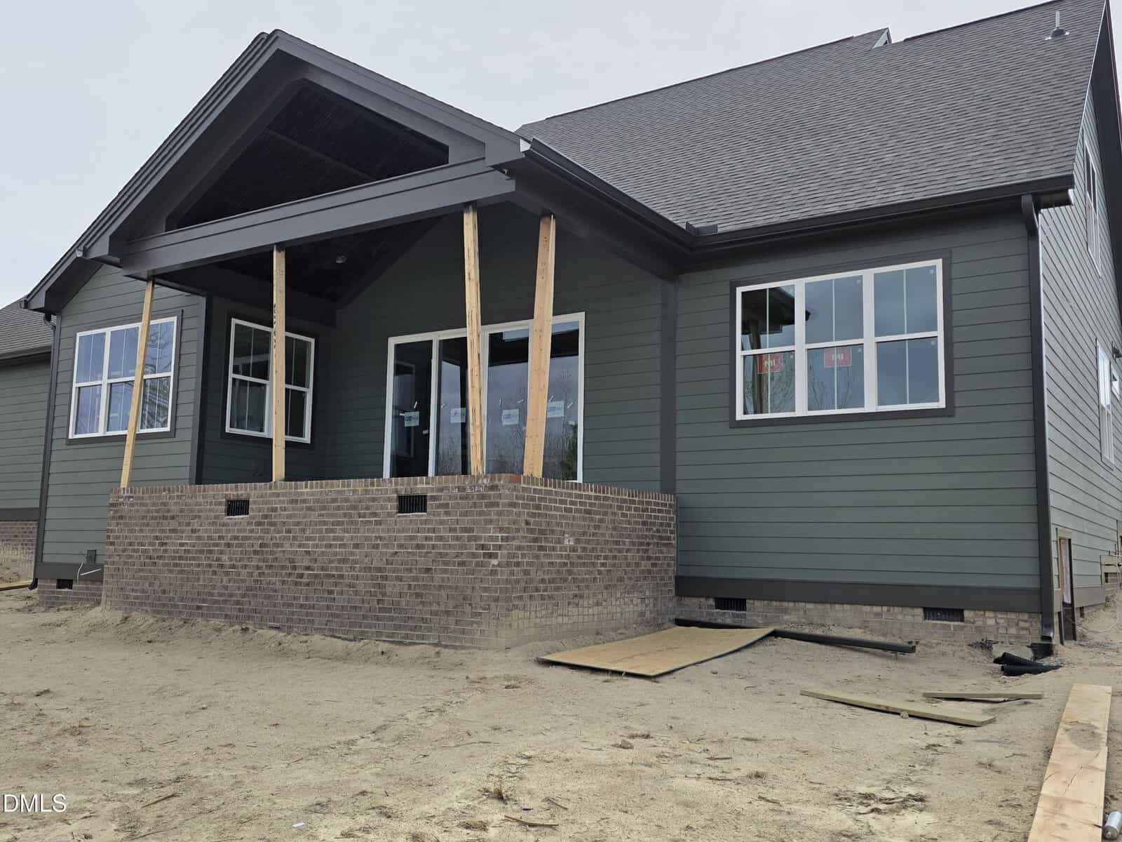 A partially constructed house with gray siding, white-trimmed windows, a covered front porch supported by temporary wooden beams, and an unfinished yard with dirt and scattered construction materials.