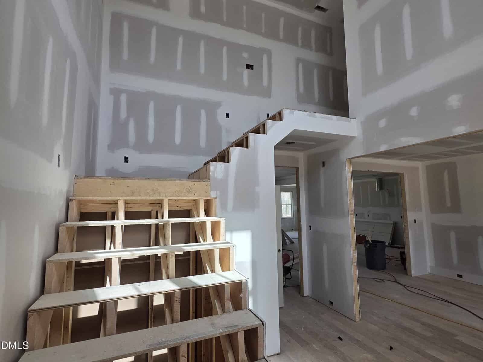 A partially constructed interior of a house features unfinished wooden stairs, exposed drywall with taped seams, and construction materials on the floor. An open area under the stairs leads to another room.