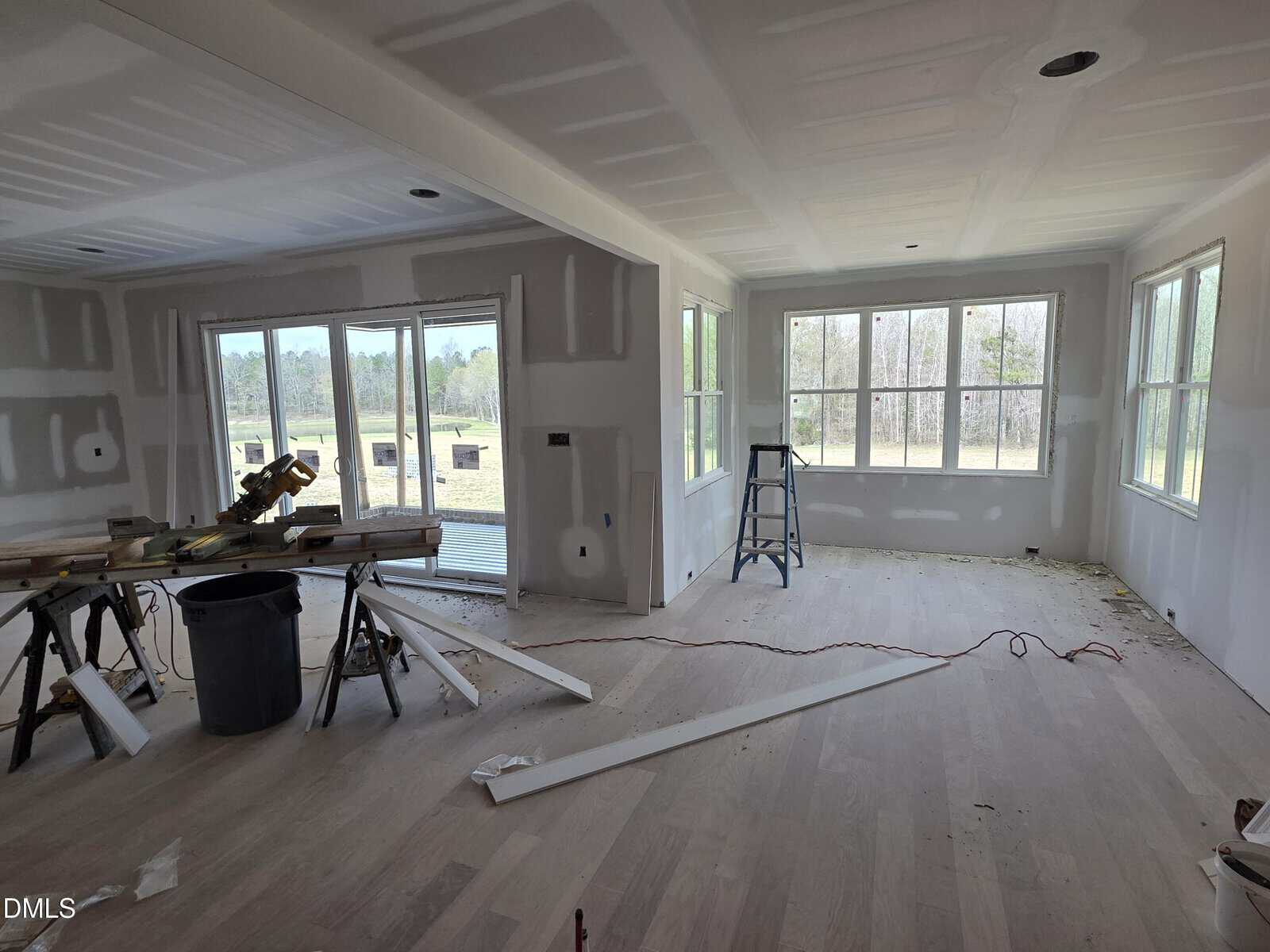 A room under construction with unfinished drywall, wooden floorboards, a ladder near large windows, tools and cut wood on a worktable, and a sliding glass door leading outside.
