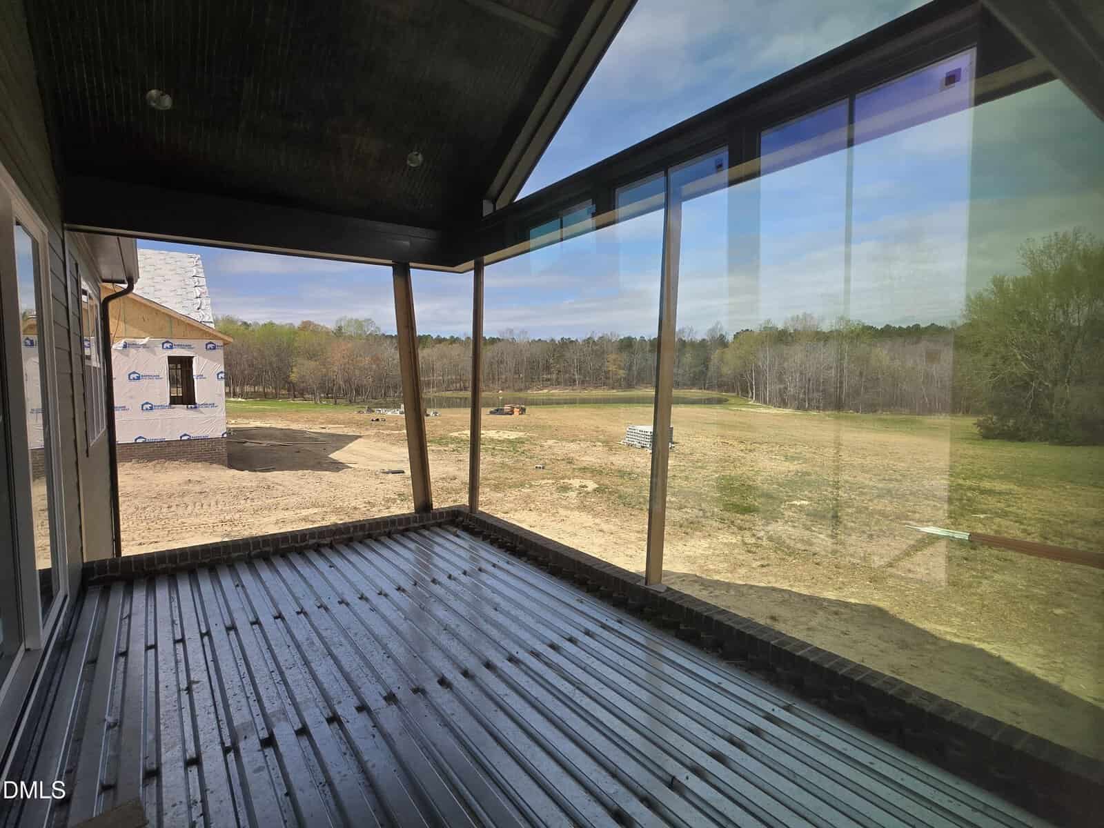 A screened-in porch with a dark wood floor overlooks a large, grassy backyard with scattered trees and construction materials. A partially built structure is visible to the left. The sky is mostly clear with some clouds.