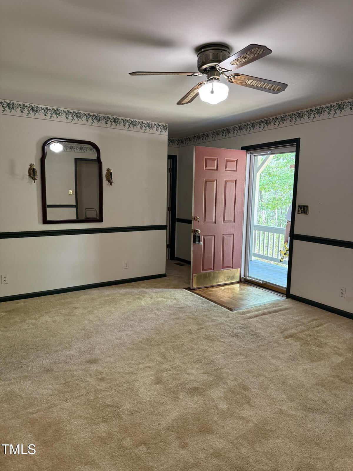 A room with beige carpet, a ceiling fan with a light, a door painted red standing open to a porch, white walls with green trim, wallpaper border, and a wall mirror with two sconces.
