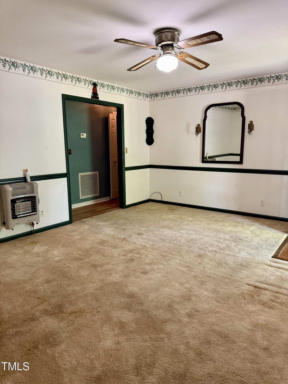 A carpeted room with white walls, green trim, a ceiling fan with a light, a wall mirror, a wall-mounted heater, and floral border near the ceiling. An open doorway leads to another area with wood flooring.