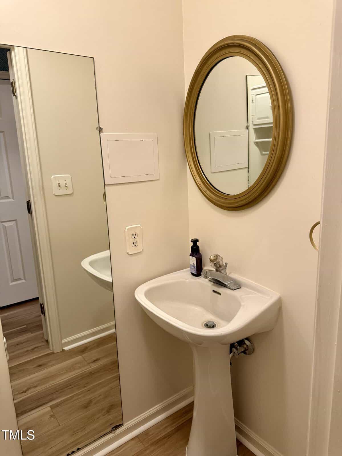 A small bathroom corner with a white pedestal sink, a soap dispenser, a gold-framed round mirror above the sink, and a tall rectangular mirror on the adjacent wall. Light wood flooring and neutral walls are visible.