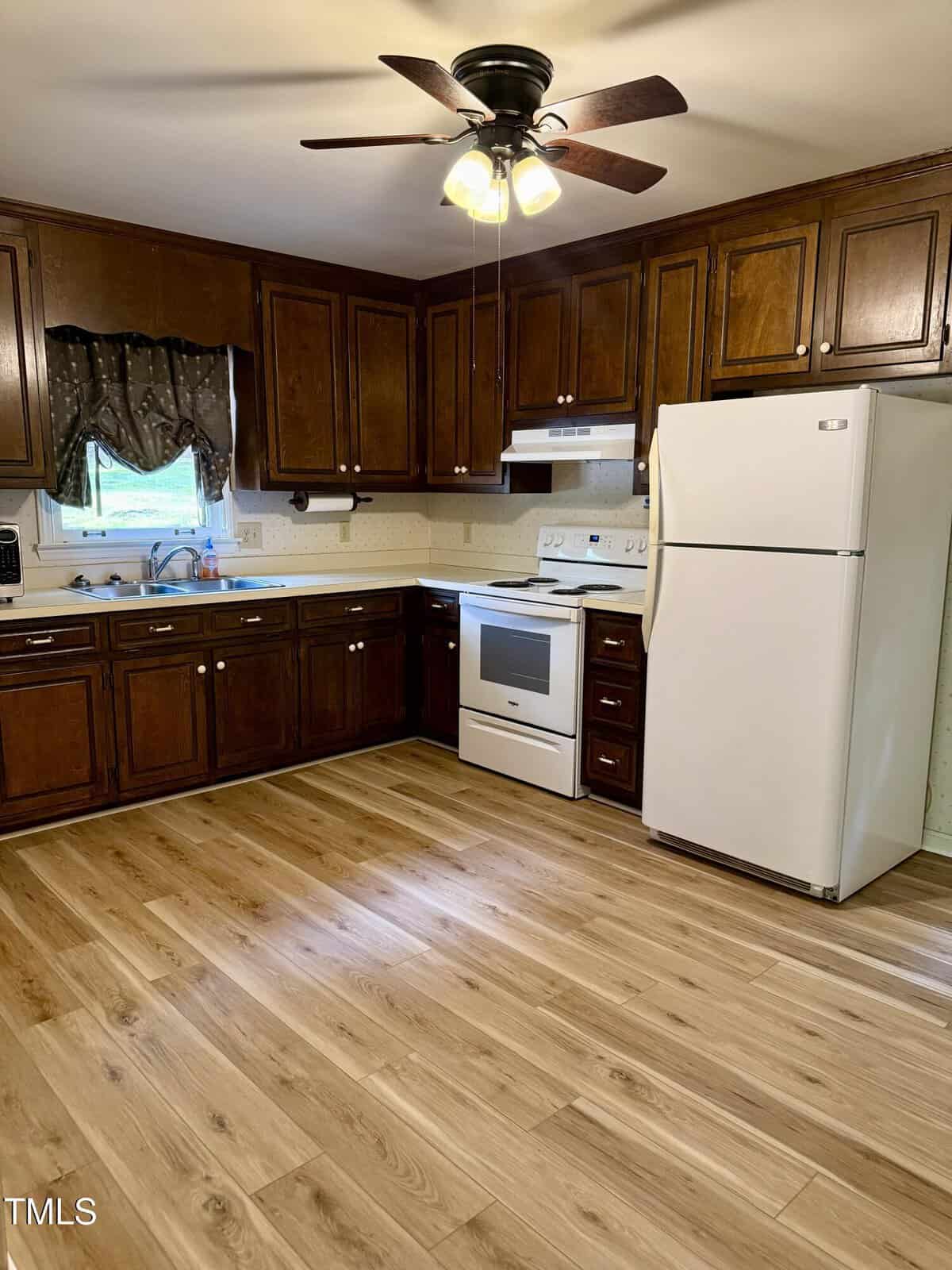 A kitchen with dark wooden cabinets, light wood flooring, white appliances including a refrigerator and stove, a ceiling fan with lights, and a small window above the sink with curtains.