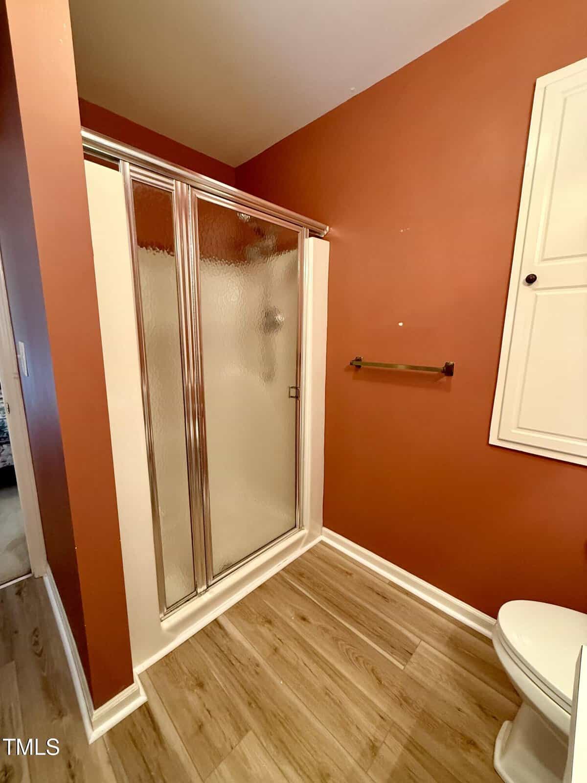 A bathroom with reddish-brown walls, a glass shower enclosure with a sliding door, a towel bar, a white wall cabinet, wood-look flooring, and a white toilet partially visible in the lower right corner.