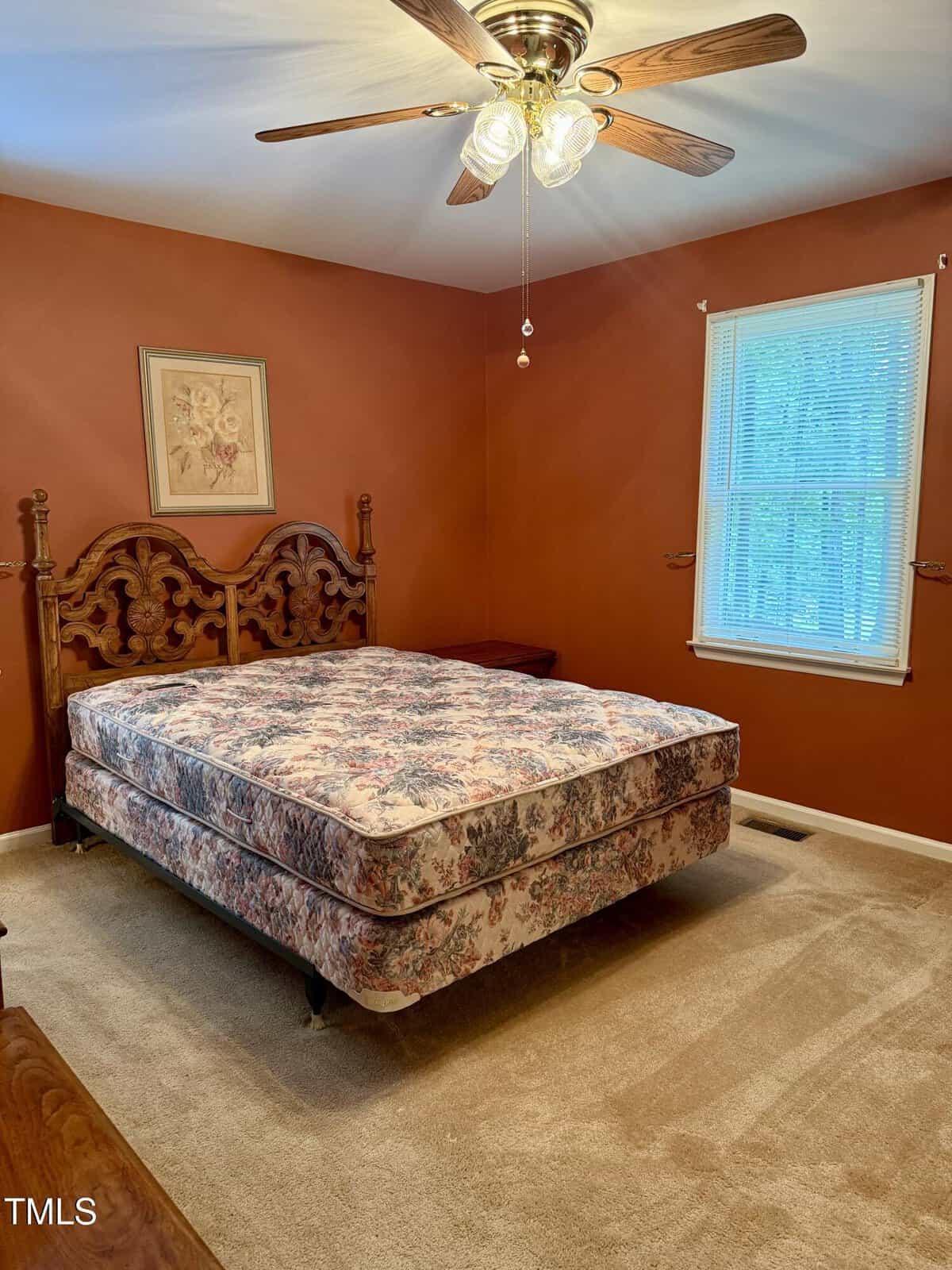 A bedroom with tan carpet, orange-brown walls, a ceiling fan with lights, a window with blinds, a wooden bed frame, and a floral-patterned mattress. A framed picture hangs above the bed.