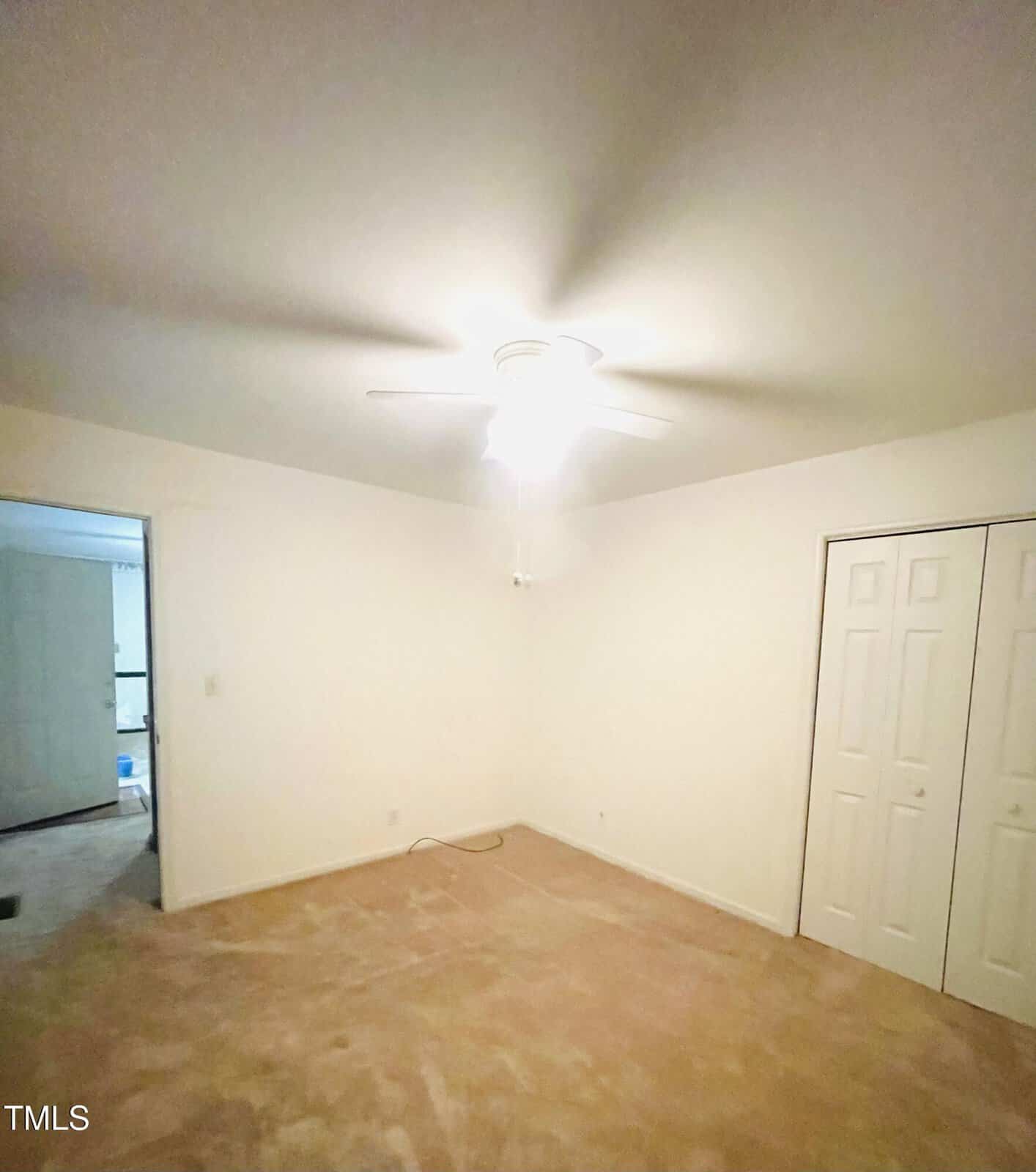 Empty room with beige carpet, white walls, a ceiling fan with light, double white closet doors, and an open doorway leading to another room. A single cord is lying on the floor.
