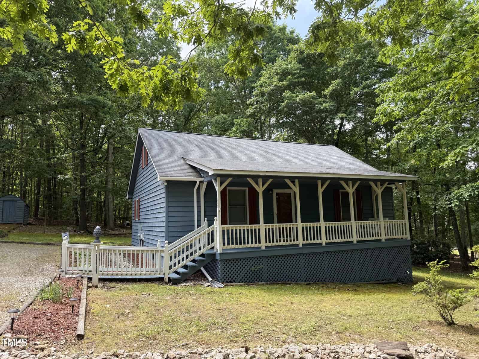 A blue, single-story house at 225 Meadow Drive in Henderson features a covered front porch with white trim, set on a grassy lot surrounded by trees. A gravel driveway runs alongside the house, with landscaped areas of mulch and rocks in the yard.