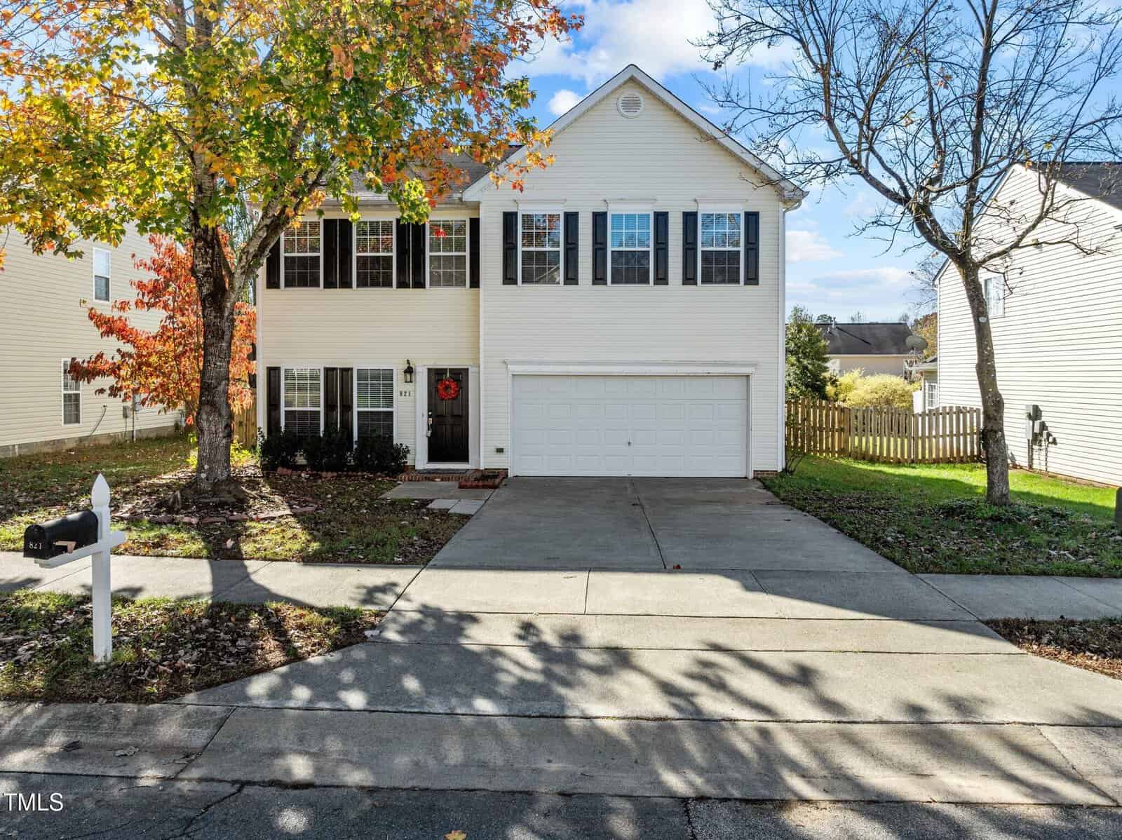 Two-story white house with black shutters, a double garage, and a concrete driveway. There are autumn trees in the front yard and a white mailbox at the edge of the driveway. The house has a simple, suburban appearance.