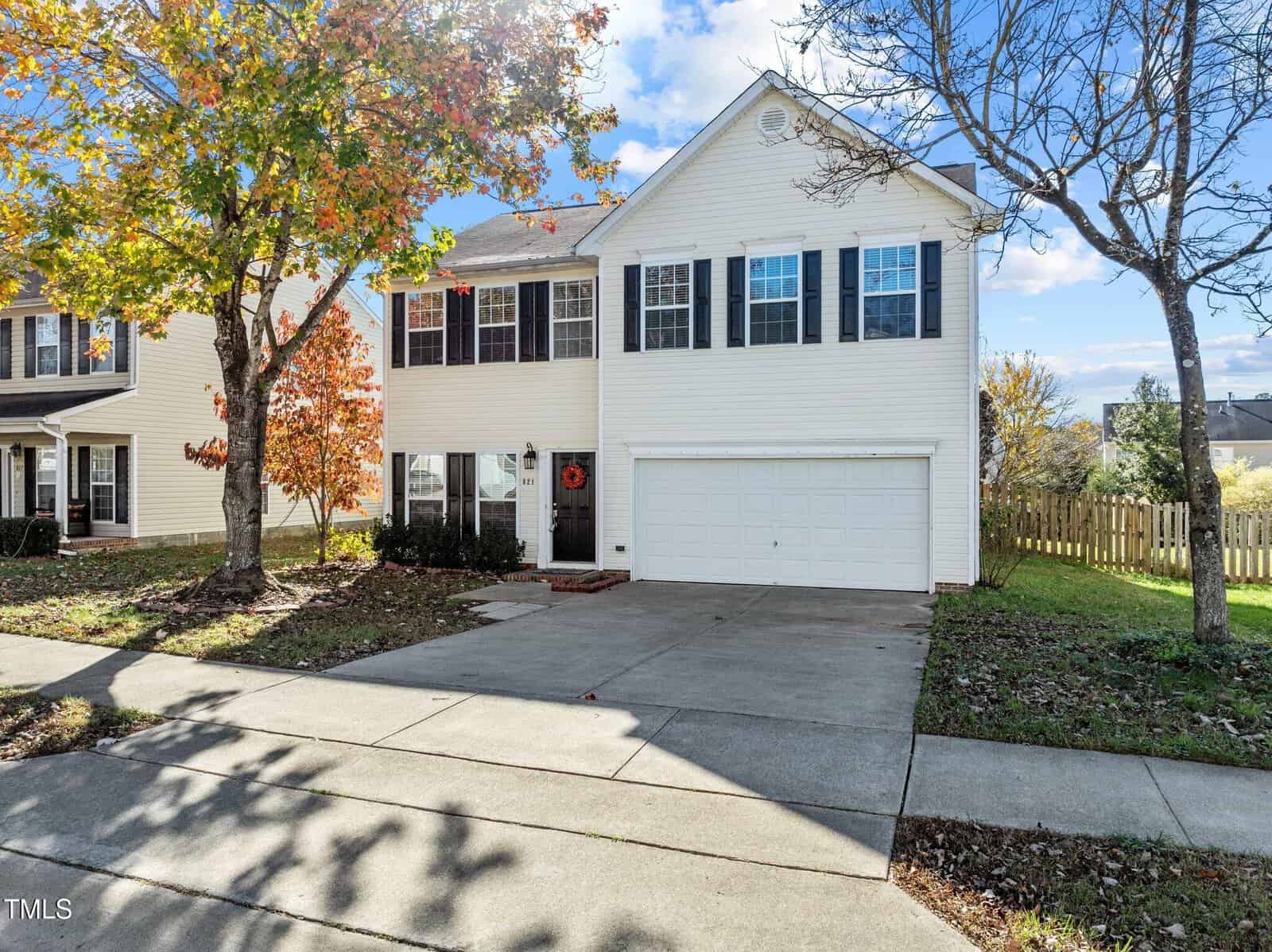 Two-story white house with black shutters, a double garage, and a concrete driveway. Autumn trees with colorful leaves frame the front yard. A wooden fence and neighboring homes are visible in the background.
