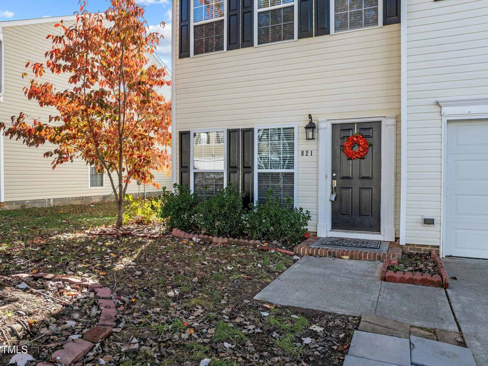 Front view of a beige two-story house with black shutters, a black front door decorated with an orange wreath, a concrete walkway, and a tree with autumn-colored leaves in the yard.