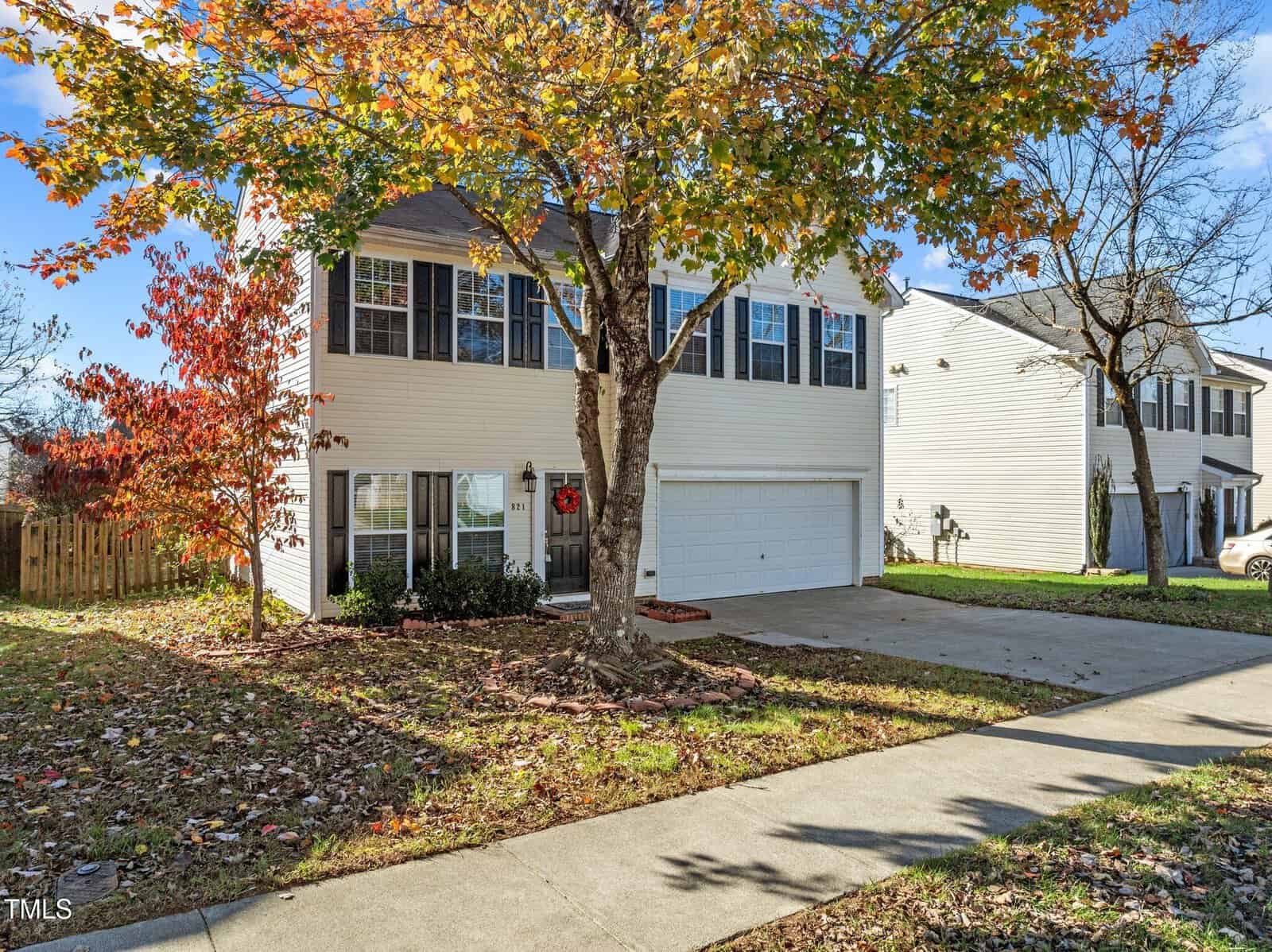 Two-story beige house with black shutters, a white double garage, and a tree with autumn leaves in the front yard. Sidewalk and driveway lead up to the house; neighboring homes are visible on both sides.
