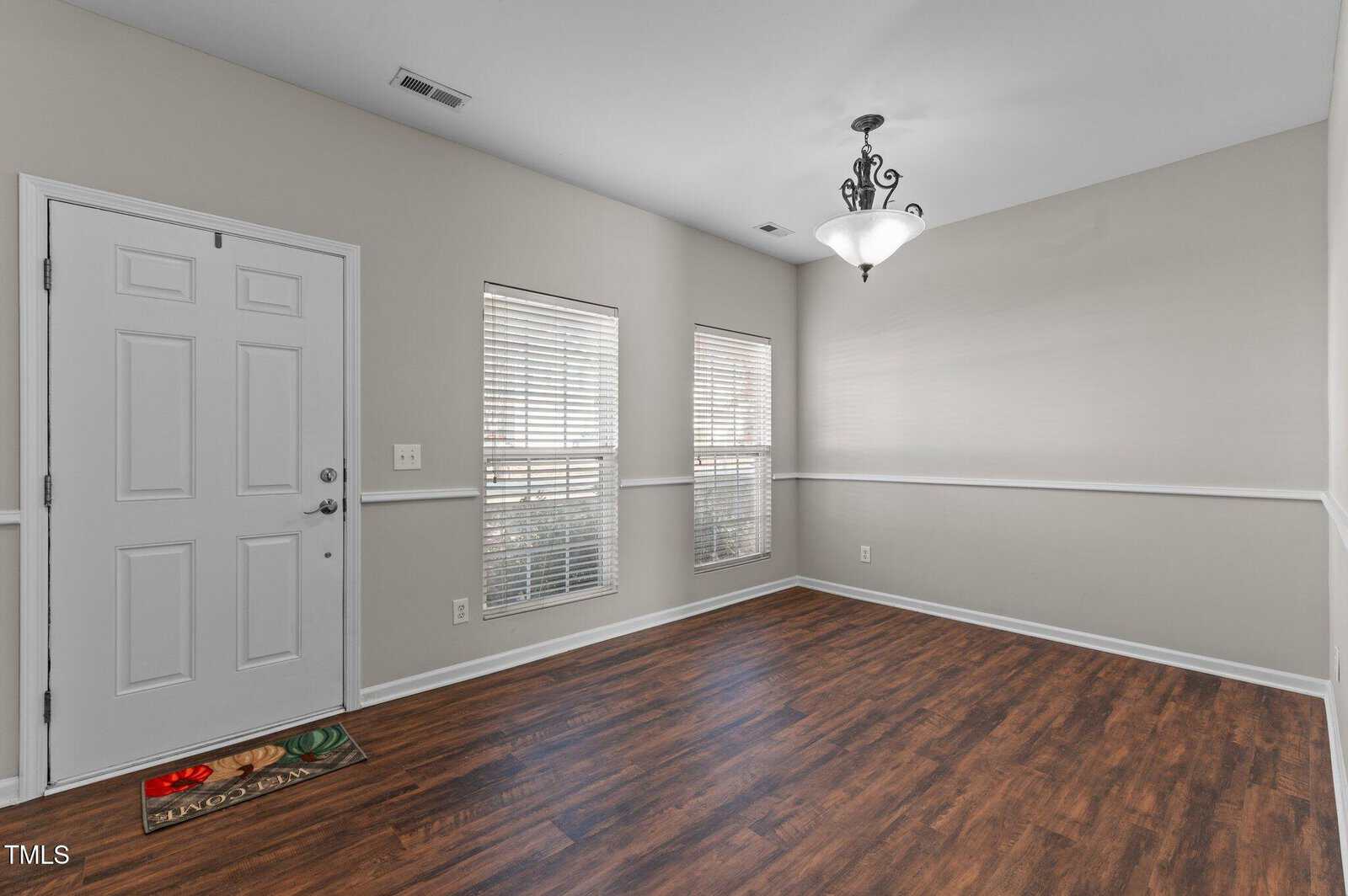 Bright, empty room with wood flooring, light gray walls, two windows with blinds, a white front door, a decorative ceiling light, and a welcome mat near the door.
