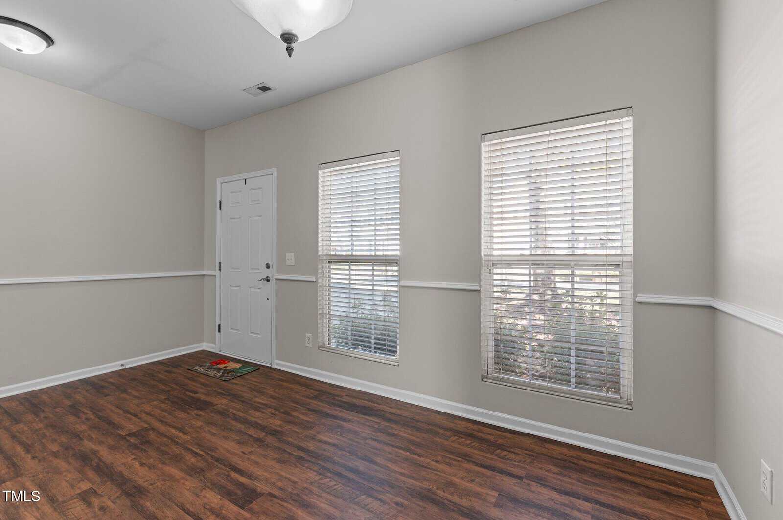 Small, empty room with beige walls, white trim, and wood flooring. Two windows with blinds are on the right wall. A white door and light fixture are also visible. There is a small welcome mat by the door.