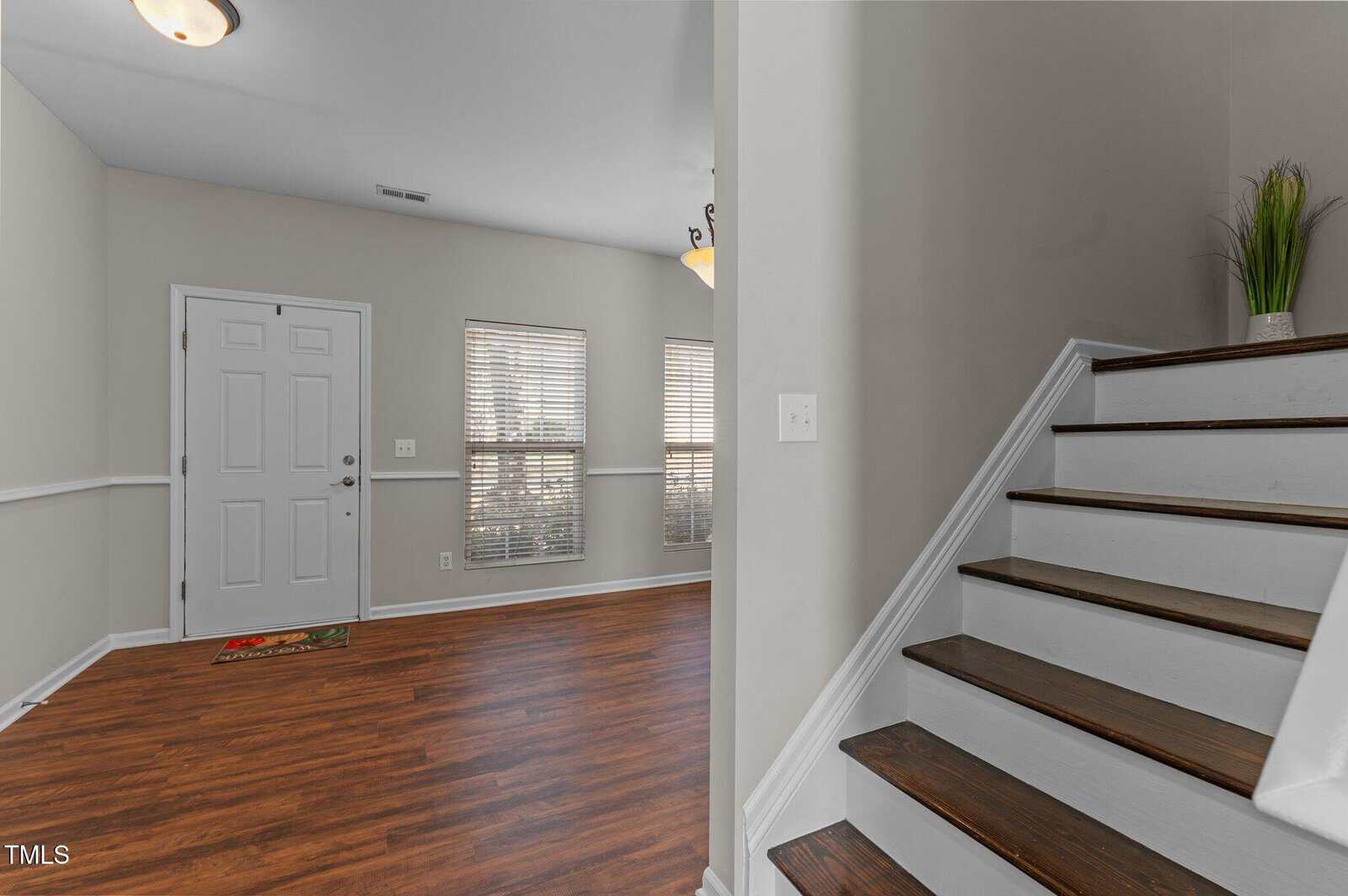 Entryway with a white front door, two windows, hardwood floors, and wooden stairs with white risers. A potted plant sits on the stair landing. Walls are painted light gray. Ceiling light fixture is visible.