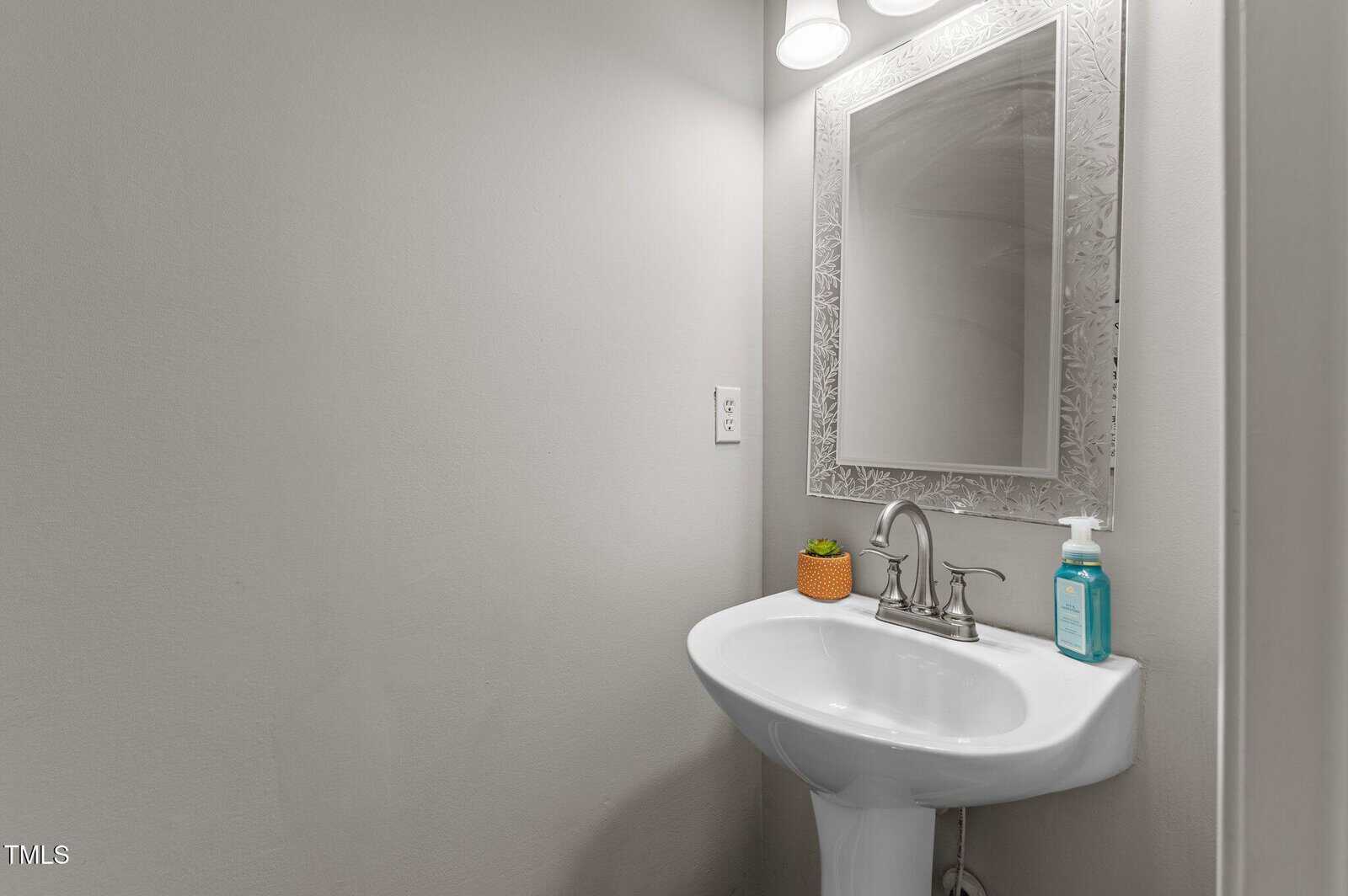 A small bathroom sink with a silver faucet, a decorative mirror above it, a small orange planter, and a bottle of soap on the side, all against a light gray wall under a pair of overhead lights.