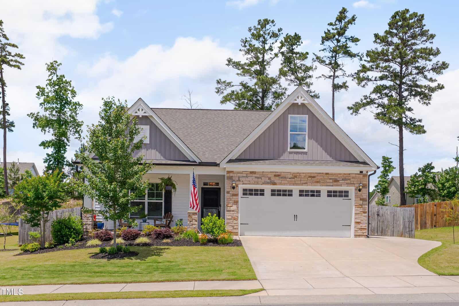 A modern suburban house with a well-kept lawn, an American flag by the front porch, a two-car garage, and stone and siding exterior, surrounded by trees and a wooden fence.
