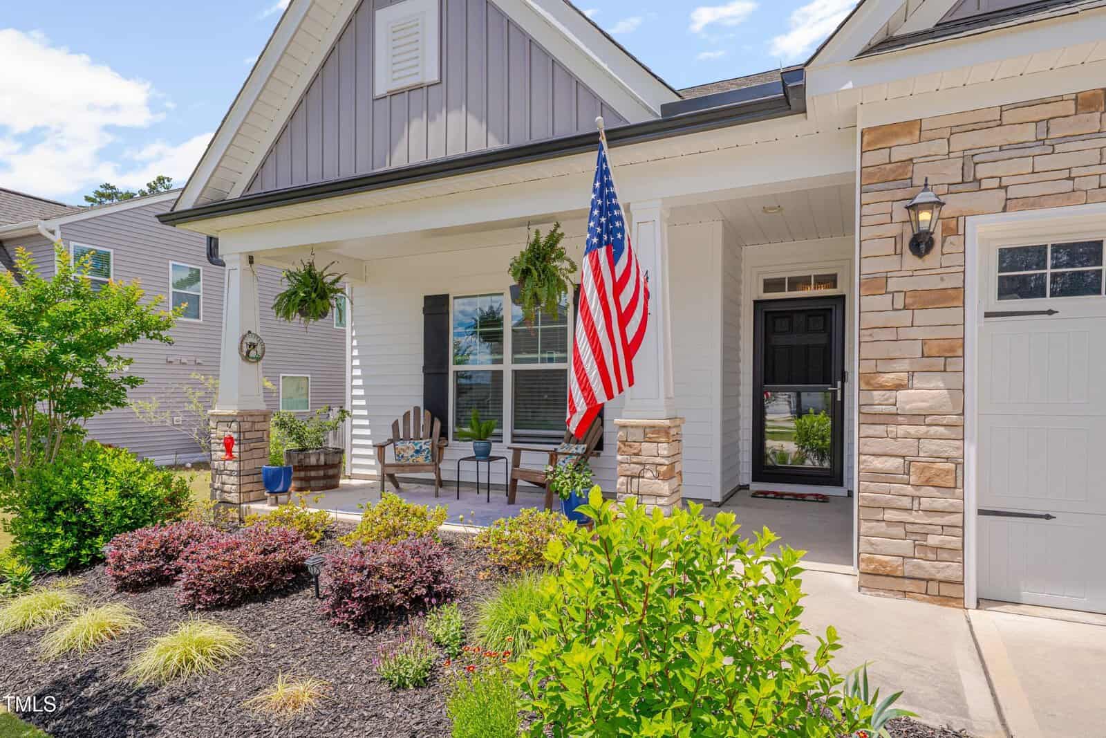 A welcoming house with a front porch featuring two chairs, hanging plants, an American flag, and stone accents. Green shrubs and bushes line the landscaped garden in front of the home.