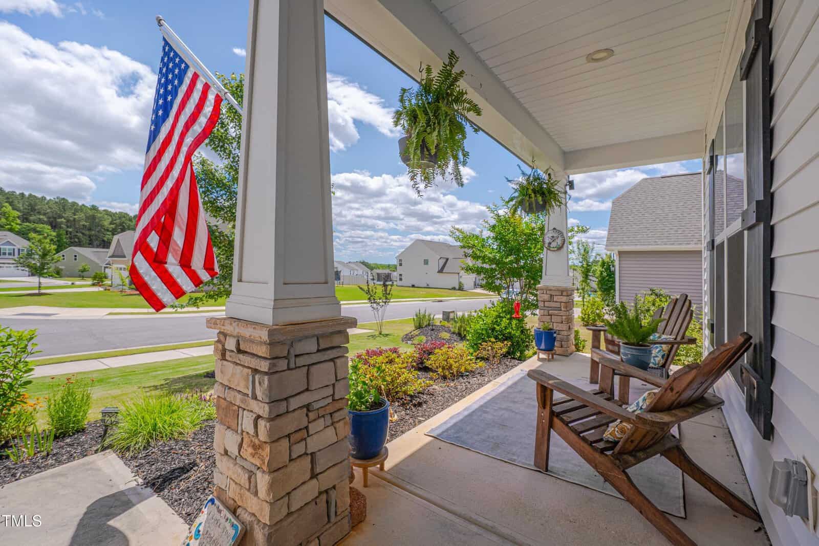 A wooden chair sits on a front porch decorated with potted plants and a large American flag. The porch overlooks a green lawn, suburban street, and neighboring houses under a bright blue sky with scattered clouds.