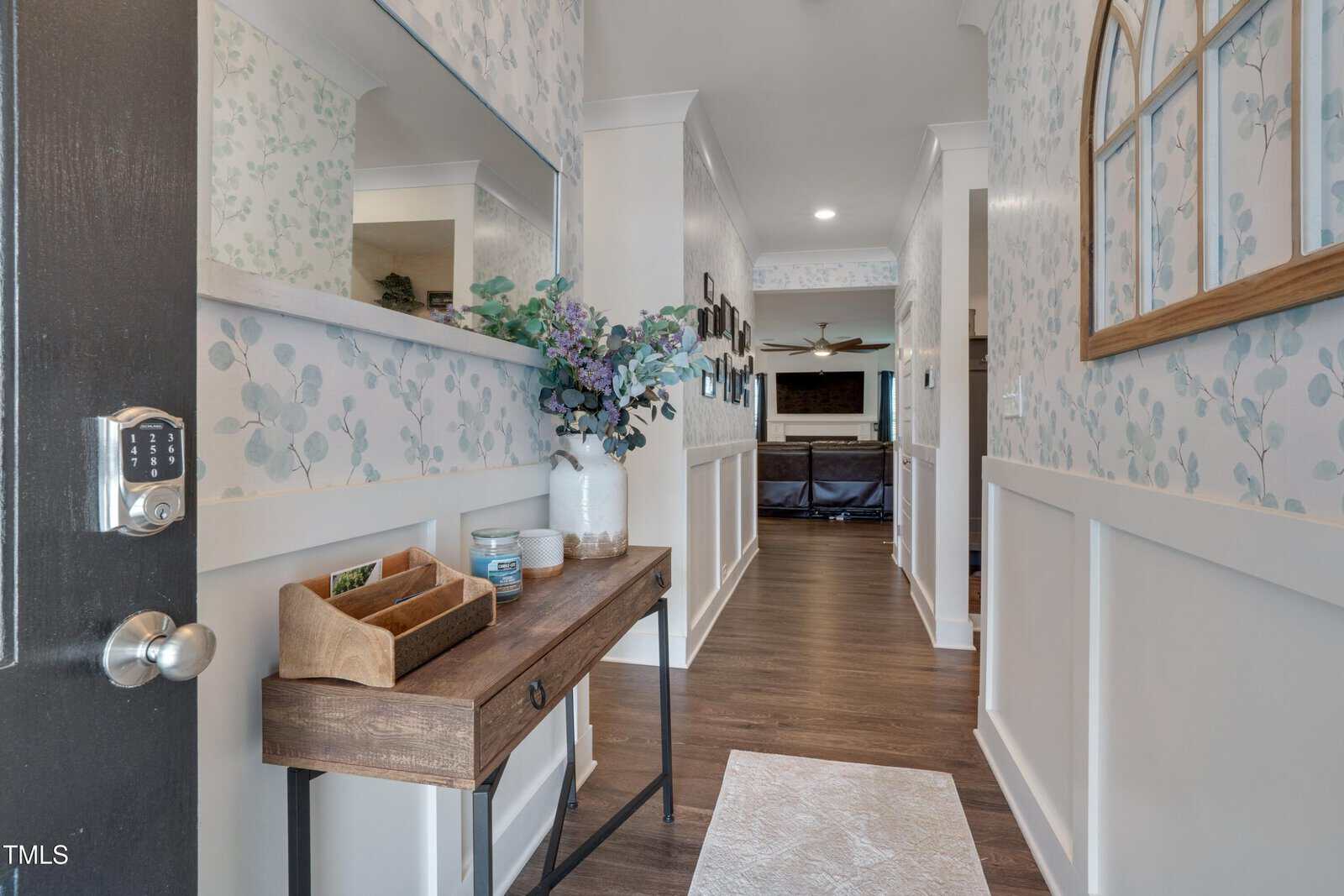 A hallway with floral wallpaper, a wooden console table holding vases, candles, and a wooden organizer, a large mirror above, and a view into a living area with a ceiling fan and TV in the background.