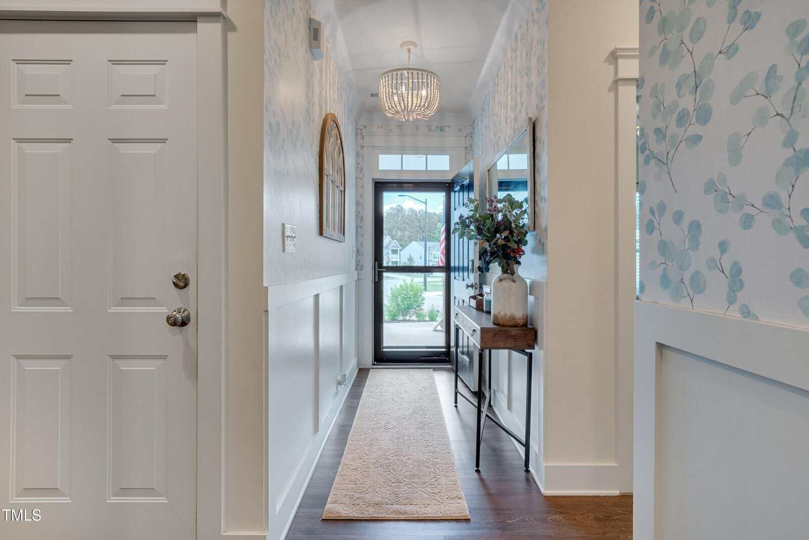 A bright, welcoming hallway with a white front door, a long beige rug on dark wood floors, floral wallpaper, a narrow console table with flowers, and a round mirror, leading to a glass-paned door with outdoor view.