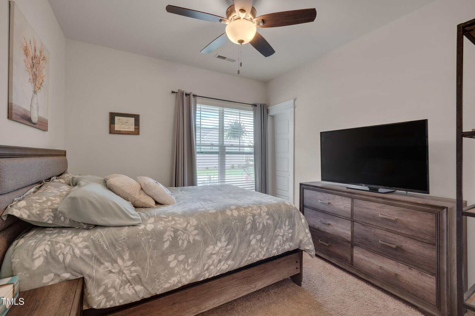 A bedroom with a double bed, floral bedding, pillows, a ceiling fan with a light, a dresser with a flat-screen TV, a window with drapes, light walls, beige carpet, and framed art above the bed and next to the window.
