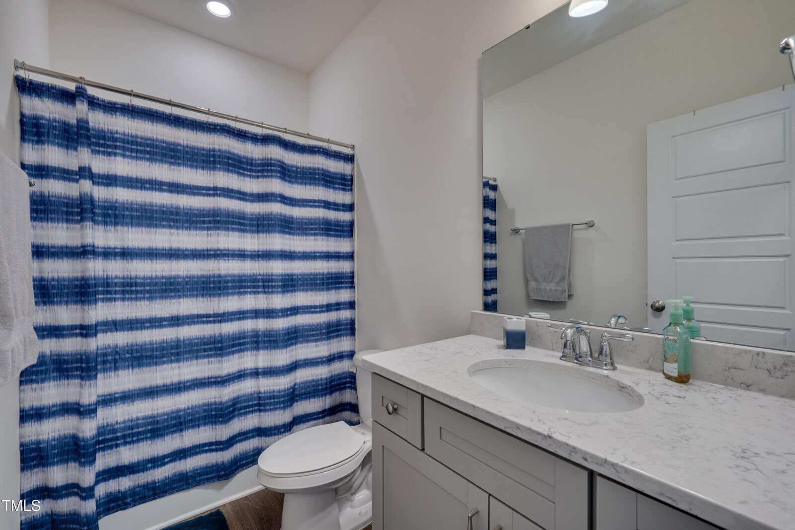 A bathroom with a white toilet, gray vanity with marble countertop, large mirror, and a shower with a blue-and-white striped curtain. A hand towel hangs on the wall; soap and toothbrush holder sit on the counter.
