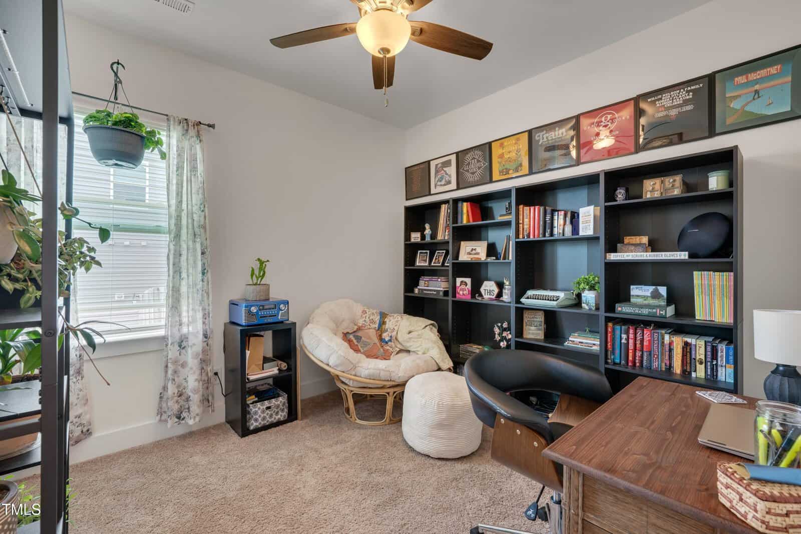 Bright home office with a ceiling fan, large bookshelf, desk, cushioned chair, window with curtain, and various plants and decorations. Record albums are displayed on top of the bookshelf. Carpeted floor and tidy appearance.