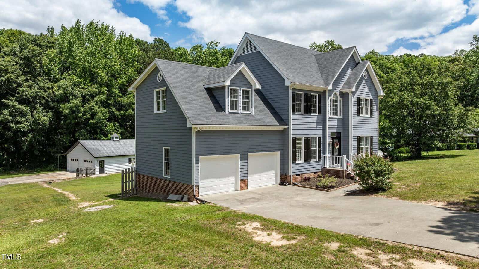 Two-story gray house with white trim and black shutters, a double garage, and a driveway. The property has a green lawn, some trees in the background, and a detached garage on the left side. The sky is partly cloudy.