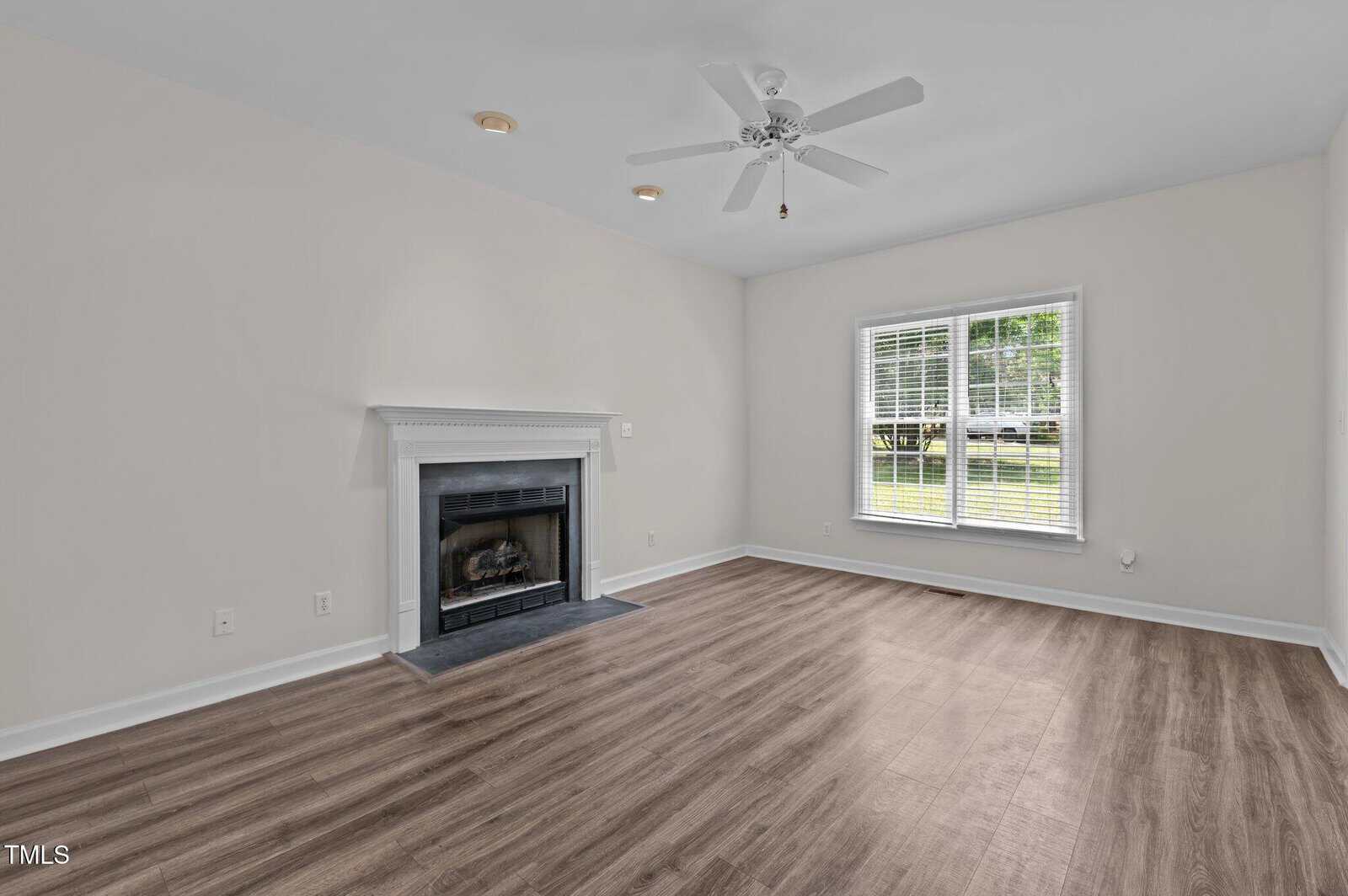 Unfurnished living room with light-colored walls, wood-look flooring, a white ceiling fan, a fireplace with a white mantel, and a large window letting in natural light.
