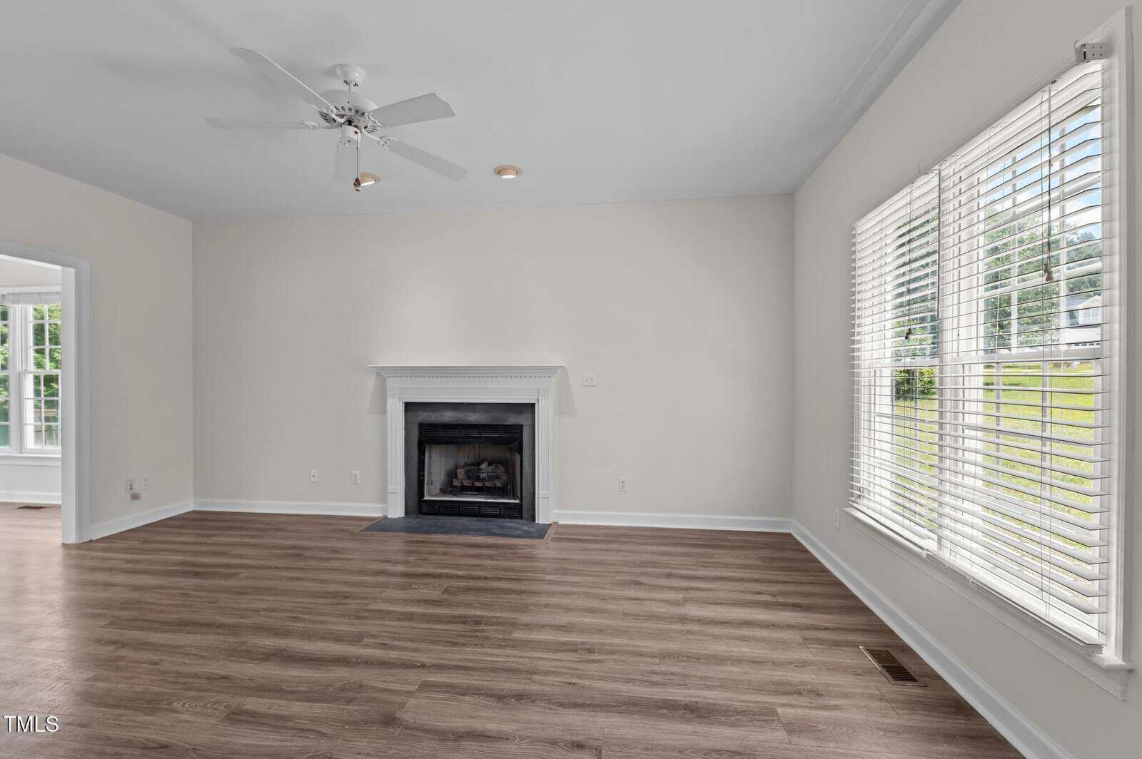 A bright living room with light-colored walls, wood flooring, a ceiling fan, a white fireplace, and large windows letting in natural light.