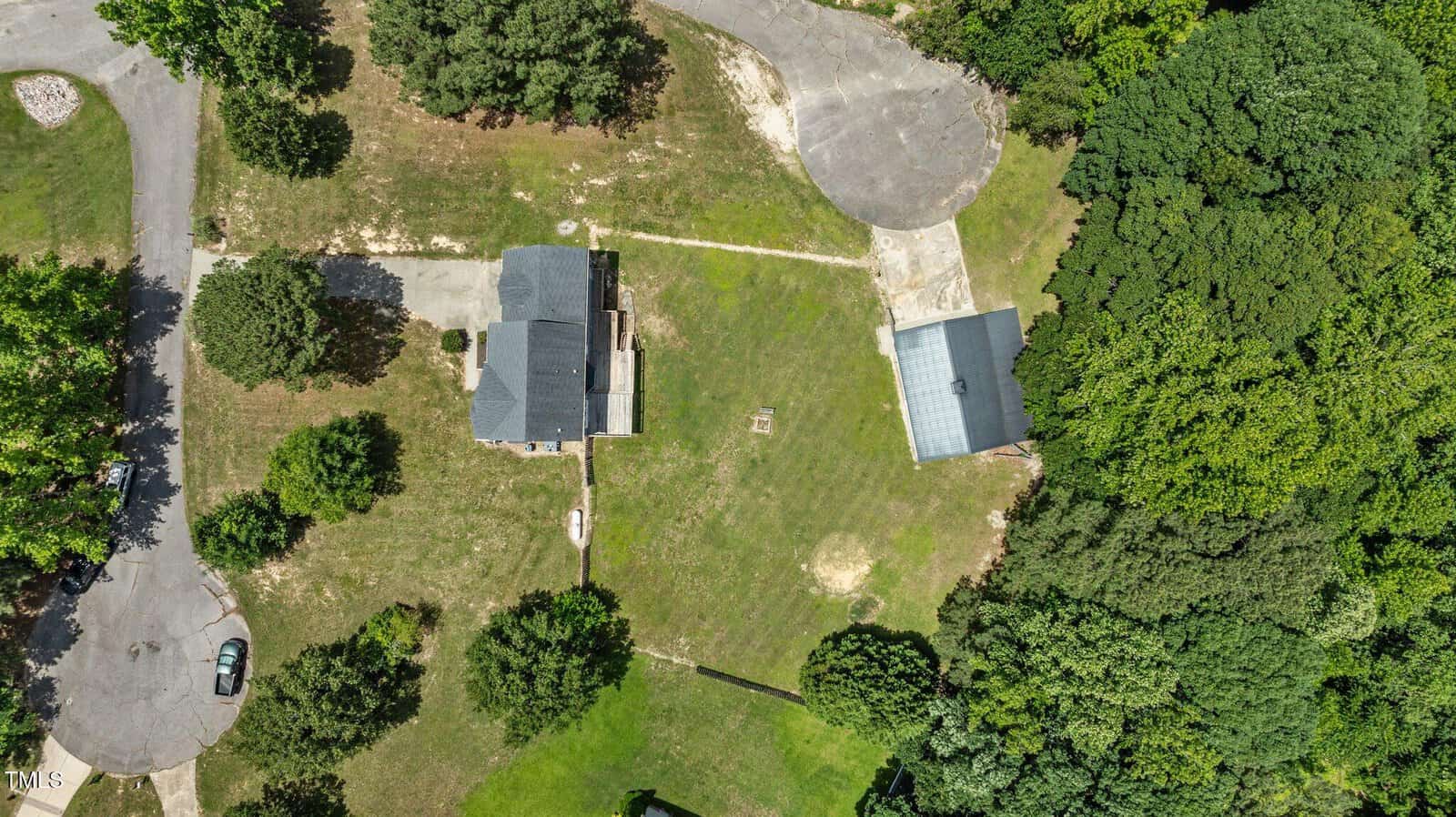 Aerial view of a house with a gray roof, a detached garage or large outbuilding, a driveway, green lawn, and several trees surrounding the property, bordered by a wooded area on one side.